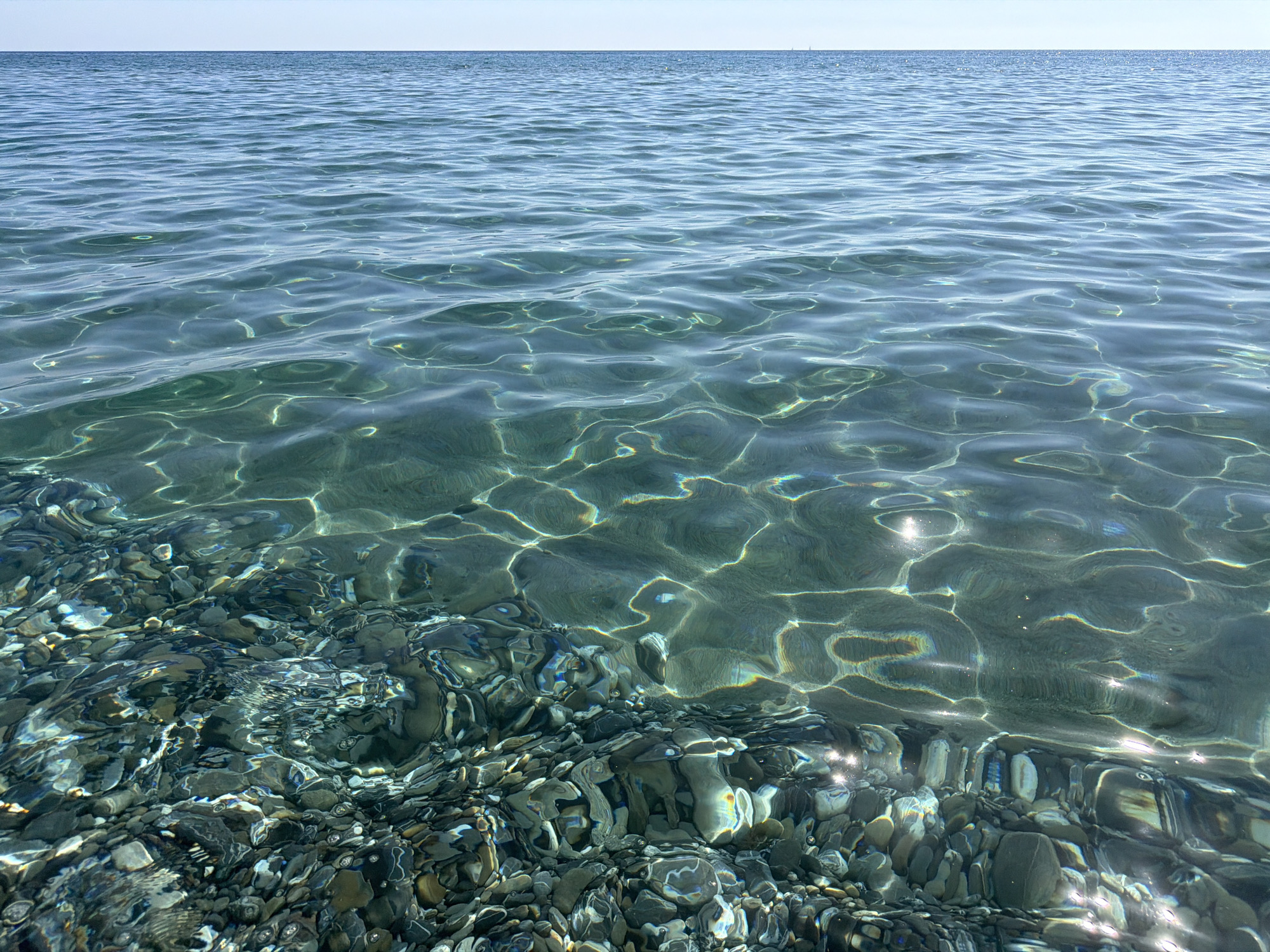 The image shows a clear, shallow sea with a rocky bottom. The water is turquoise and translucent, allowing visibility of the pebbles and stones beneath. Sunlight refracts on the surface and through the water, creating patterns of light and shadow. The horizon is visible as a straight line in the upper portion of the image. The scene is likely located at the beach in Ascea, Italy, given the presence of characteristic stones and the typical sea color of the Cilento coast. There are no people visible in the image.