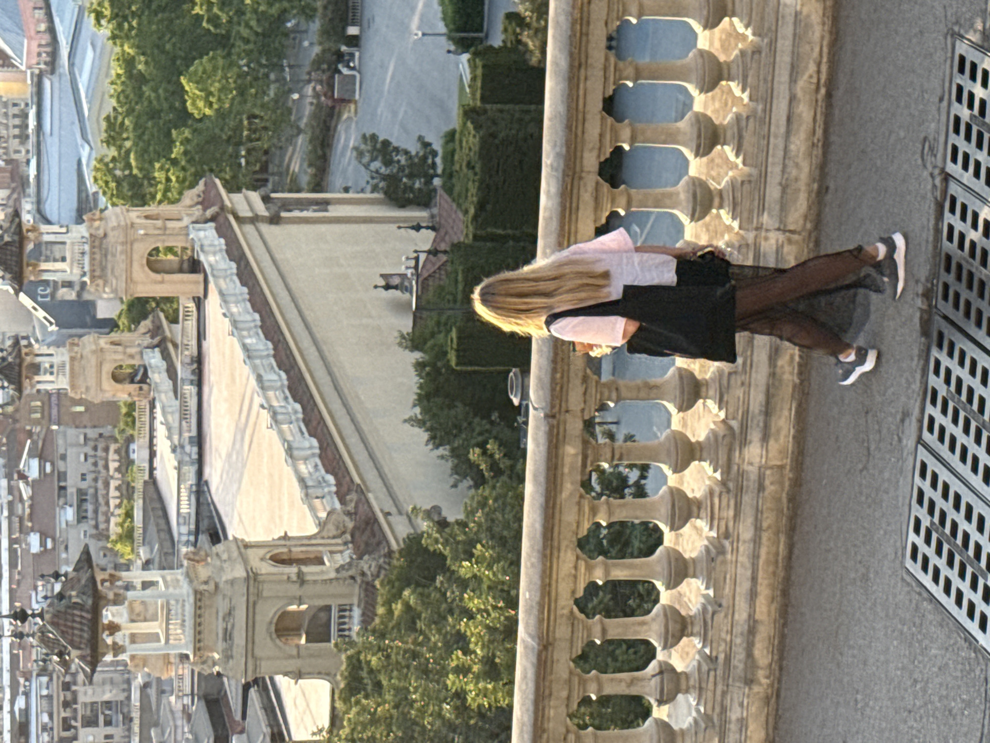 The image shows a young woman with blonde hair walking away from the camera on a paved walkway in front of a decorative stone railing. She is wearing a light-colored t-shirt, black sheer leggings, and sneakers, and carrying a black tote bag. Behind her, a sprawling cityscape with buildings and trees is visible under a bright sky. The architectural style of the buildings, particularly the ornate towers with tiled roofs and arched windows, suggests the location is Barcelona, Spain, likely from a vantage point overlooking the city. The sunlight casts long shadows, indicating it might be late afternoon or early morning. A metal grate is visible in the foreground.