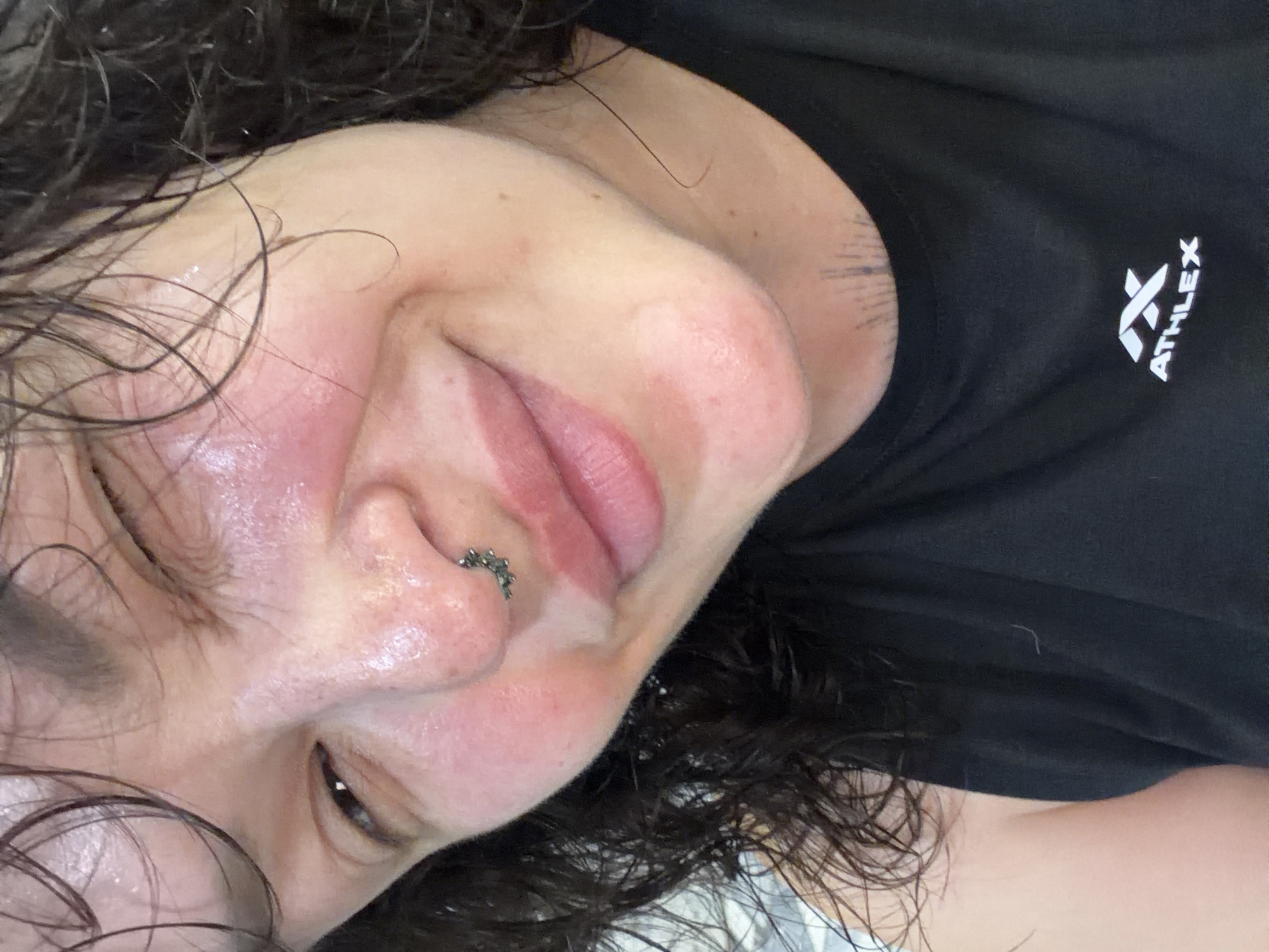 The image is a close-up, slightly angled selfie of a person with dark, wet hair. The person is wearing a black tank top with a white logo that reads "ATHLEX". They are winking and smiling, with a nose ring visible. Their skin appears flushed and slightly sweaty, and there is a tattoo visible on their neck. The background is indistinct, appearing to be a light-colored wall or surface. The location in the city of Chaikovsky, Russia, is not determinable from the provided image.