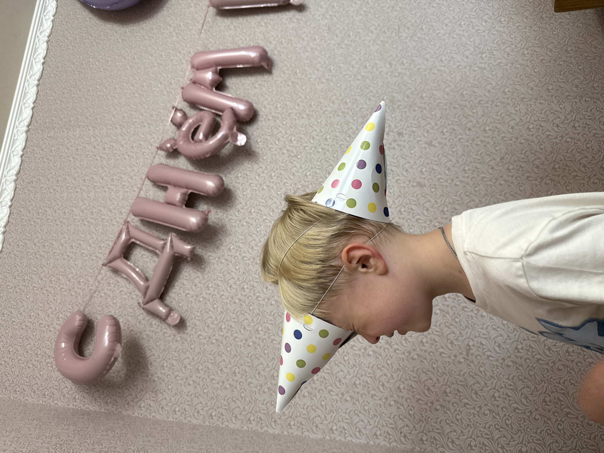In the city of Luzhkovo, Russia, a young boy with blonde hair is pictured in profile, wearing a polka-dotted party hat. The hat is positioned at an odd angle, with one side covering his eye and the other extending out behind his head. He is wearing a white t-shirt and a thin chain necklace. In the background, a string of pink balloon letters spells out a Russian phrase, possibly a birthday greeting. The wall behind the boy and the balloons is covered in a textured wallpaper with a subtle pattern. A hint of another balloon, purple in color, is visible on the far right. The scene appears to be indoors, likely in a home setting.