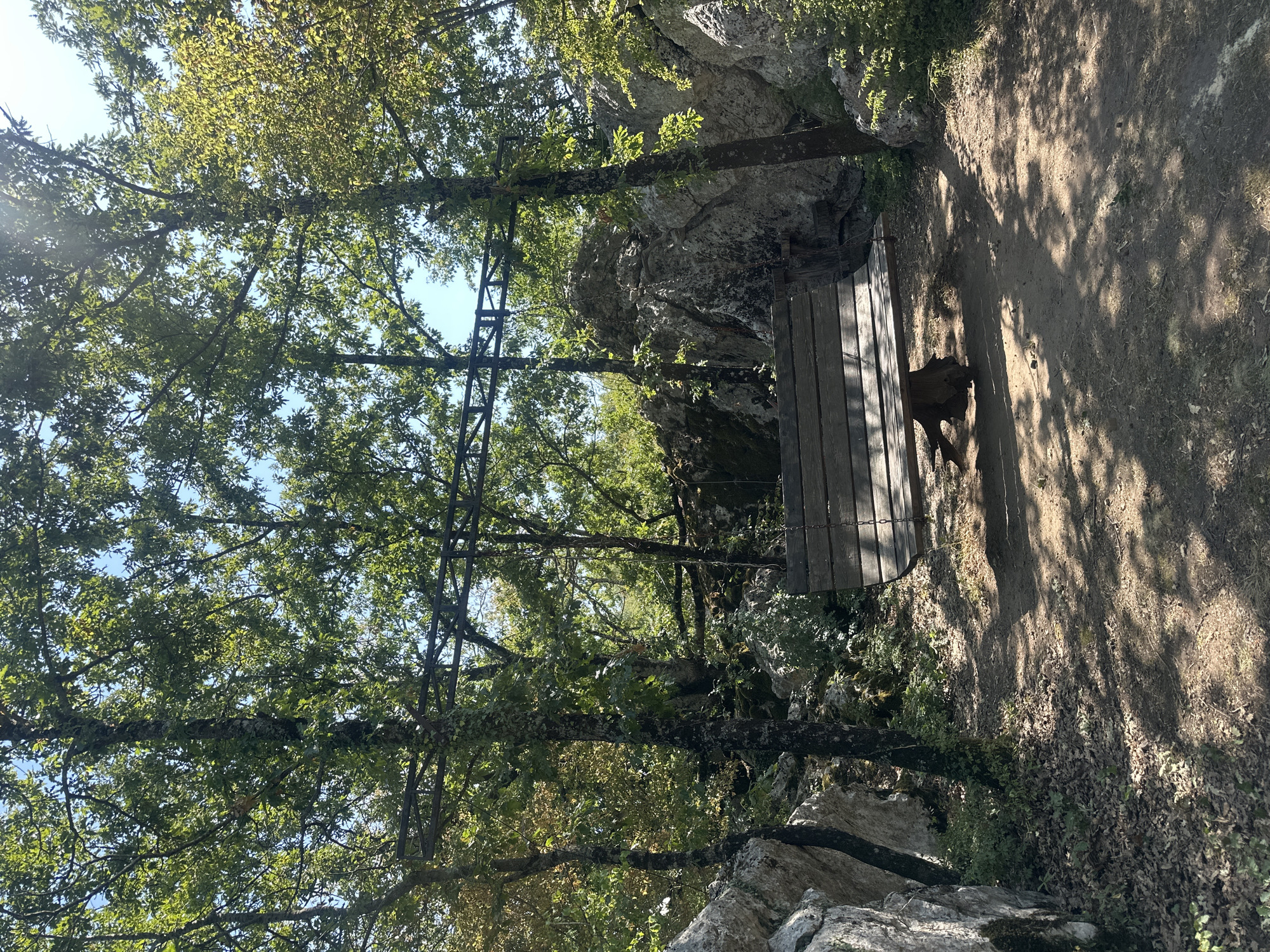 The image shows a wooden swing hanging from a metal frame supported by two trees. The swing is in a clearing surrounded by rocks and trees. The ground is covered in dirt and fallen leaves. Sunlight filters through the leaves, casting dappled shadows on the ground. The scene is set in a natural environment, likely a forest or park. The location is identified as Bukovik, Montenegro. There are no people visible in the image.