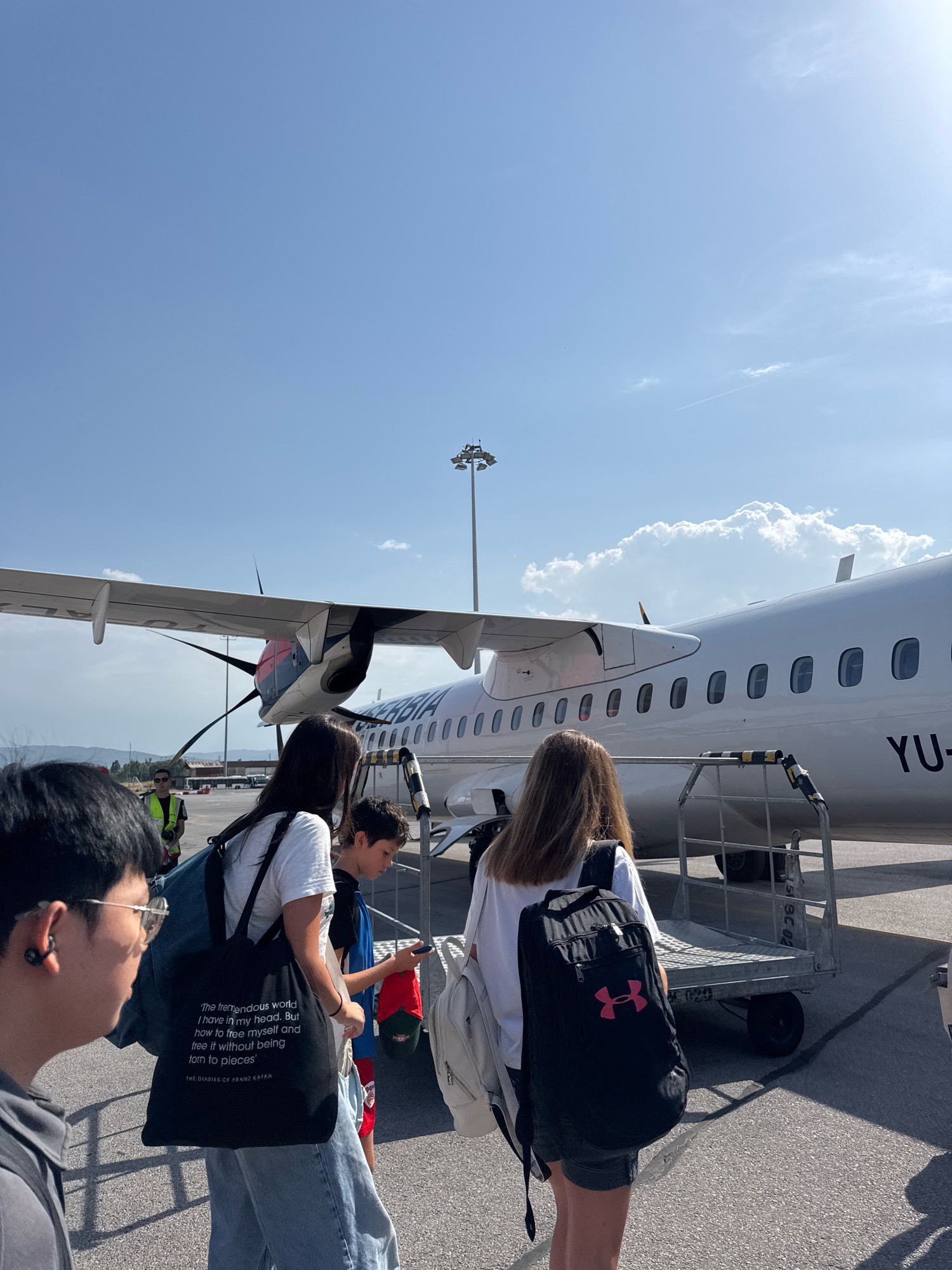 This is a vertical, eye-level shot of several people boarding a small passenger airplane. The airplane is white with a red and blue tail section and the text "SERBIA" is visible on its side. A propeller is visible on the left wing. People are walking towards the plane, some are carrying backpacks. The environment appears to be an airport tarmac under a clear blue sky with some clouds. The location is the city of Sofia, Bulgaria.