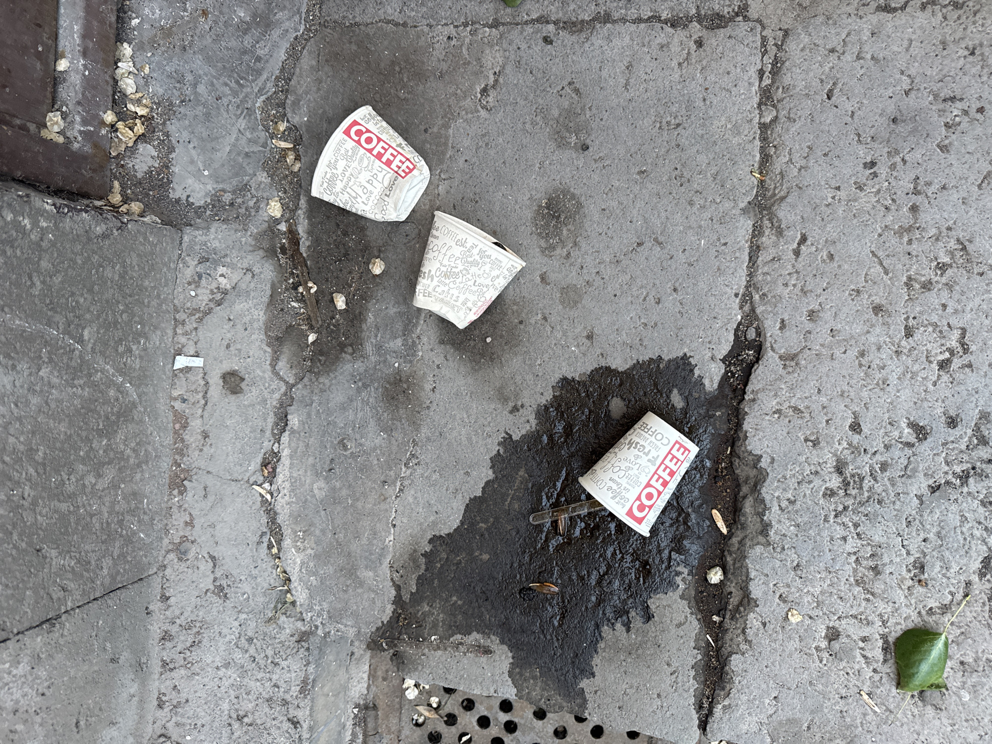 The scene is a section of Yerevan, Armenia's, street, likely a sidewalk, showing three discarded paper coffee cups, their red "Coffee" labels prominent. One cup lies amidst a dark, spilled coffee stain, its plastic stirrer nearby. The sidewalk is constructed of uneven, gray concrete slabs. Additional debris includes scattered bits of leaves, twigs, and other litter. A storm drain is visible in the bottom of the frame.