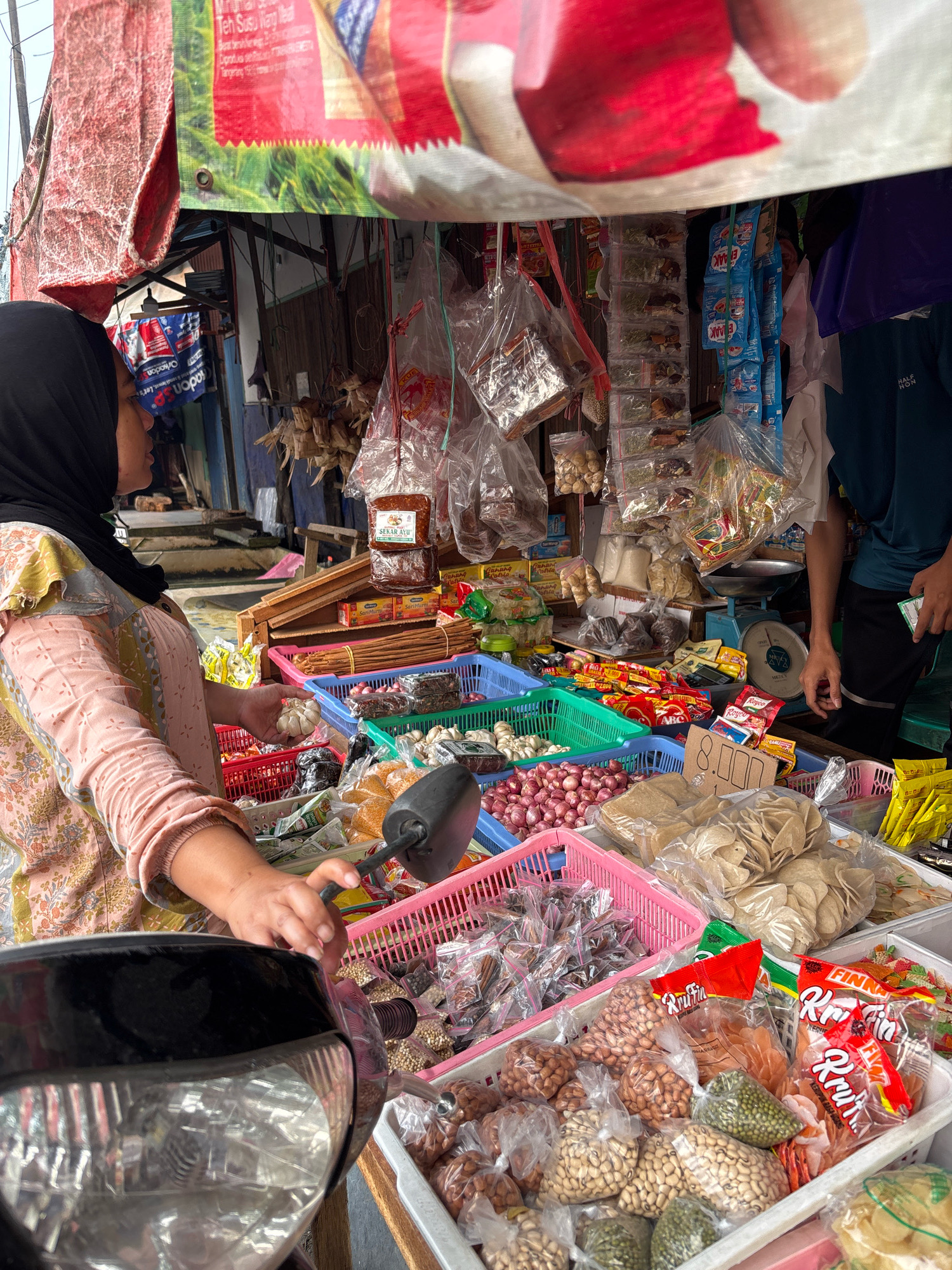 This is a medium-shot, outdoor photograph of a street food stall in Banjarmasin, Indonesia. The stall is packed with various food items, including produce, packaged snacks, and dried goods. In the foreground, a woman wearing a black hijab and a patterned shirt is seen from the side, reaching into a basket of garlic. She is partially obscured by the front of a motorcycle. The stall is covered by a red and white striped tarp. Behind the woman, shelves are laden with more products, and a blue scale sits on a table. The overall impression is one of a bustling, vibrant market scene.