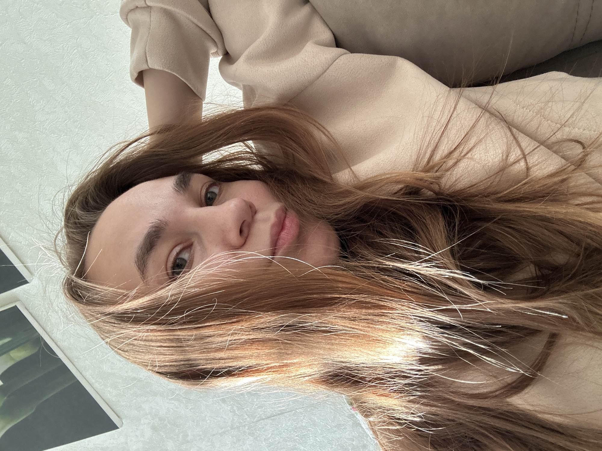 A young woman is depicted in a close-up, high-angle shot, appearing to be lying on her back and looking directly at the camera. The entire image is tilted diagonally. She has long, light brown hair, which is spread out around her head and partially covers her face, with individual strands catching the light. Her eyes are open, and her lips form a gentle, soft smile, conveying a calm expression.
She is wearing a light-colored, possibly beige or cream, long-sleeved garment that appears soft and comfortable, possibly a sweater or a jacket. This clothing item is prominently featured on the upper and right sides of the frame. The background is an indoor setting, likely in Uva, Russia, showing a light-colored wall with a subtle textured finish. On the left, two framed items, possibly photographs or minimalist artwork with dark and light elements, are visible on the wall. The lighting suggests natural daylight, creating bright highlights in her hair. There is no visible text in the image.