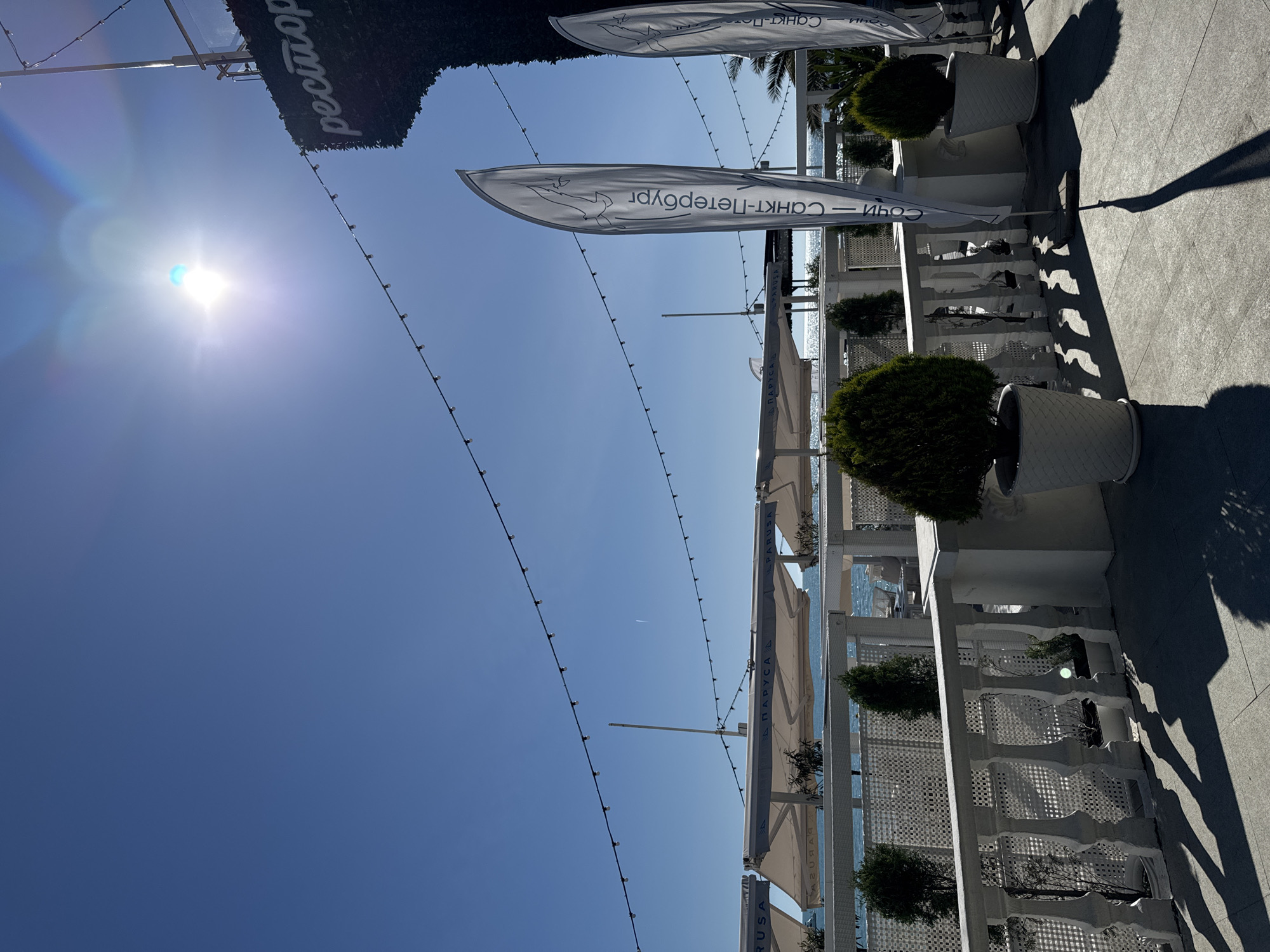 The image captures a bright, sunny outdoor restaurant or cafe terrace alongside the sea, likely in Sochi, Russia. The scene is dominated by white architectural elements, including decorative balustrades or fences, and white planters holding small, manicured green bushes.
Above, tall, narrow flags or banners billow, displaying text. One flag clearly reads "САНКТ-ПЕТЕРБУРГ" (Saint Petersburg) and features a dolphin or sea creature logo. Another flag segment bears the word "СОЧИ" (Sochi), directly confirming the location context. Below these, beige or light-colored awnings provide shade over unseen seating areas, one of which shows the text "ПАРУСА" (Sails). A dark green sign, possibly made of artificial foliage, hangs above, inscribed with "Ресторан" (Restaurant) in white letters.
Overhead, strings of small lights are suspended, likely for decorative evening illumination. The ground is paved with light grey tiles, where long, distinct shadows are cast by the structures, indicating strong direct sunlight. The sky is a clear, vibrant blue, with a prominent sun and lens flare in the upper left portion of the frame. A faint airplane contrail is visible in the distance against the blue sky. The background reveals the sparkling blue expanse of the sea.
No people are visible in the tranquil scene. The time of day appears to be late morning or early afternoon, given the bright sun and shadow length, on a perfectly clear and warm weather day.