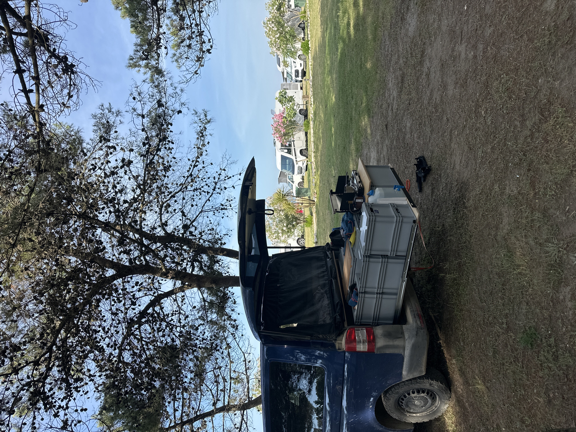 The image shows the rear of a blue van parked on a grassy area, possibly a campsite, in Donji Štoj, Montenegro. The van's rear door is open, revealing what appears to be storage or camping equipment inside, including gray storage boxes. A tree with numerous pine cones is visible to the left of the van. In the background, there are several white recreational vehicles (RVs) or campervans parked on the grass. The sky is blue with a few clouds. The ground around the van consists of grass and dirt. A black object that appears to be camera equipment is also on the ground near the rear of the van.