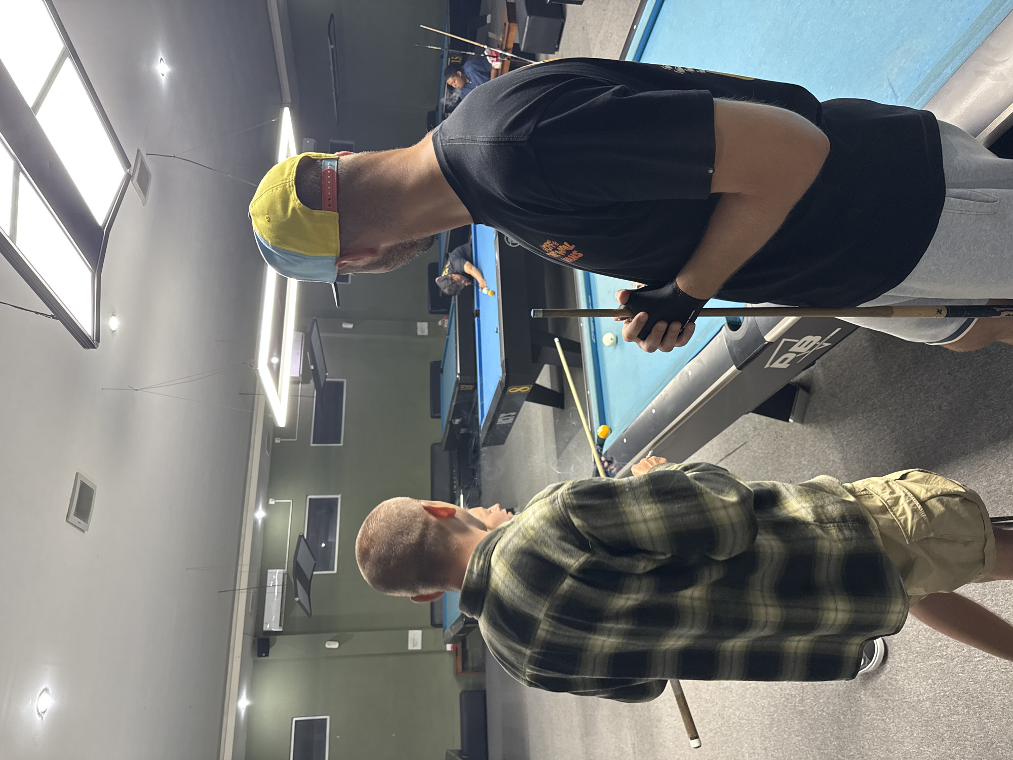 The image depicts an indoor pool hall, likely located in Kuta Selatan, Indonesia. In the foreground, two men stand near a bright blue pool table. The man on the left is wearing a green and black plaid shirt and tan shorts, with his back to the camera. The man on the right, also with his back to the camera, wears a black t-shirt and grey shorts, and a yellow and blue baseball cap. He holds a pool cue in his right hand, gloved with black fingerless gloves.
Further in the background, other pool tables are visible. On one table, a man in a black shirt is actively playing pool, positioned over the table with a cue. Another person, wearing a dark blue shirt, is seated nearby. The environment suggests a recreational setting, with typical pool hall lighting and seating. The walls are painted a dark green, and the floor is carpeted in grey.
