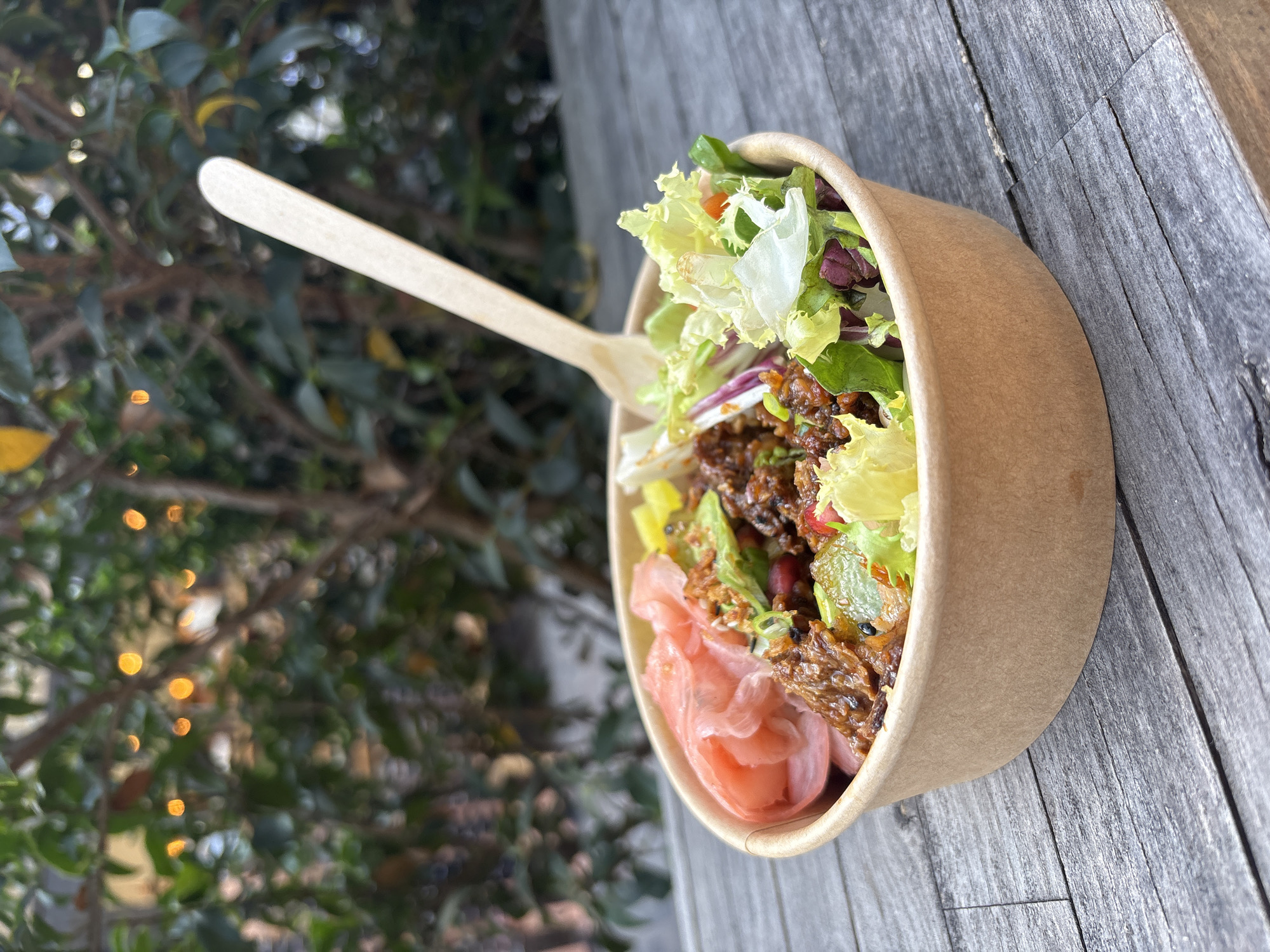 A high-angle medium shot depicts a food bowl with a disposable wooden fork resting on a wooden table outdoors. The bowl contains a salad with mixed greens, pickled ginger, and what appears to be cooked meat or a meat substitute. The bowl is made of brown paper material. The table is made of light gray wood and is placed outside, next to a bush with small yellow lights. The image lacks identifying information beyond these elements, preventing a specific determination of the location in London, United Kingdom.
