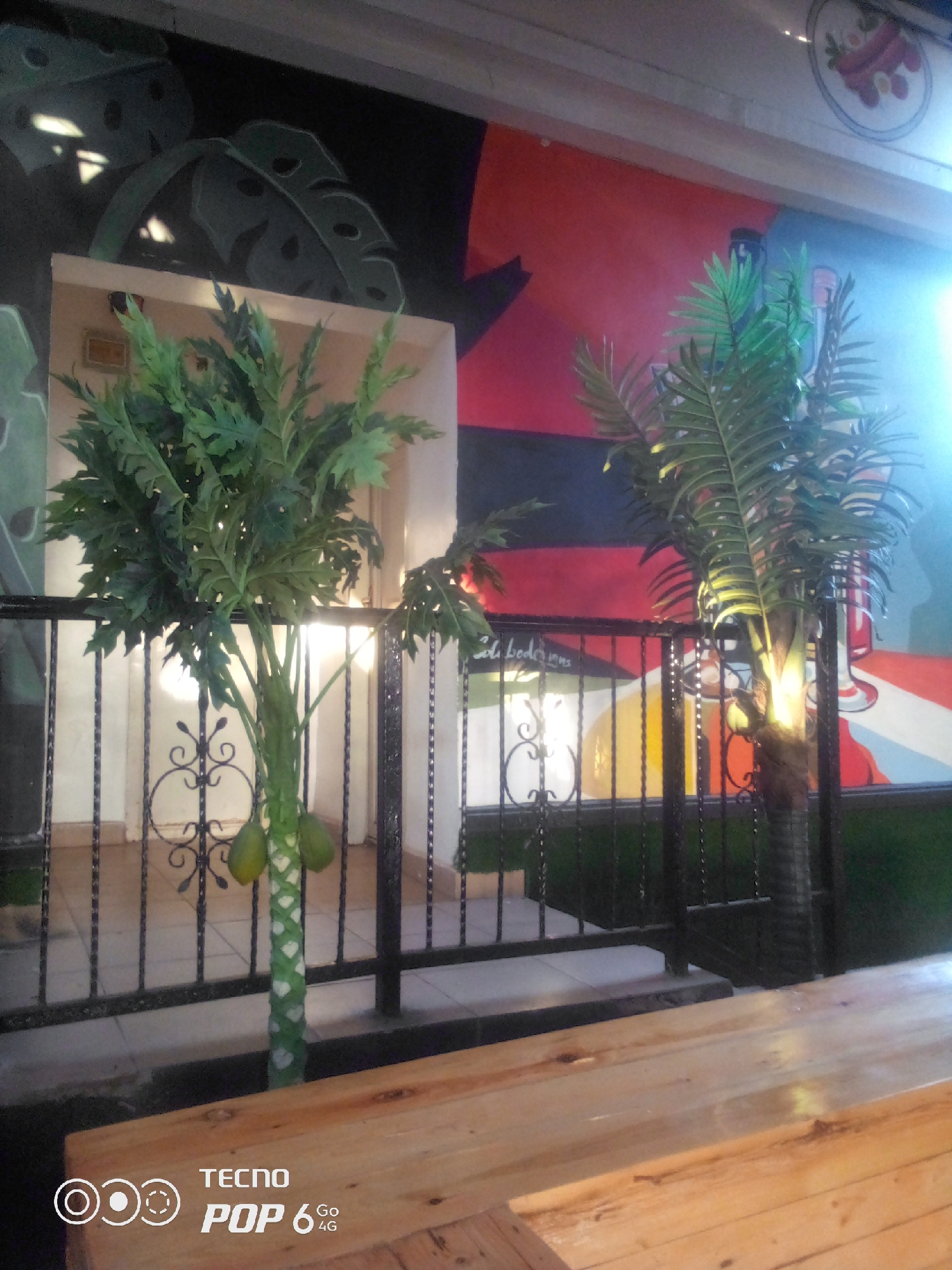 The image captures an outdoor seating area, likely a cafe or restaurant, with a rustic wooden table in the foreground. Behind the table, a black wrought-iron fence encloses a paved area with two artificial potted plants. The plant on the left resembles a papaya tree with green artificial fruit, while the one on the right is a palm tree. The background features a vibrant mural painted on a wall, depicting stylized leaves and abstract shapes in shades of black, green, red, and yellow. The mural has a bright, artistic feel. A small sign with handwritten text is visible on the wall behind the papaya tree. The overall ambiance suggests a casual, possibly tropical-themed establishment. No people are visible in the scene. The presence of the "TECNO POP 6 4G" watermark at the bottom left indicates the image was taken with a Tecno Pop 6 smartphone. The description of the location as Howayi, Nigeria, is provided without supporting visual evidence within the image itself.
