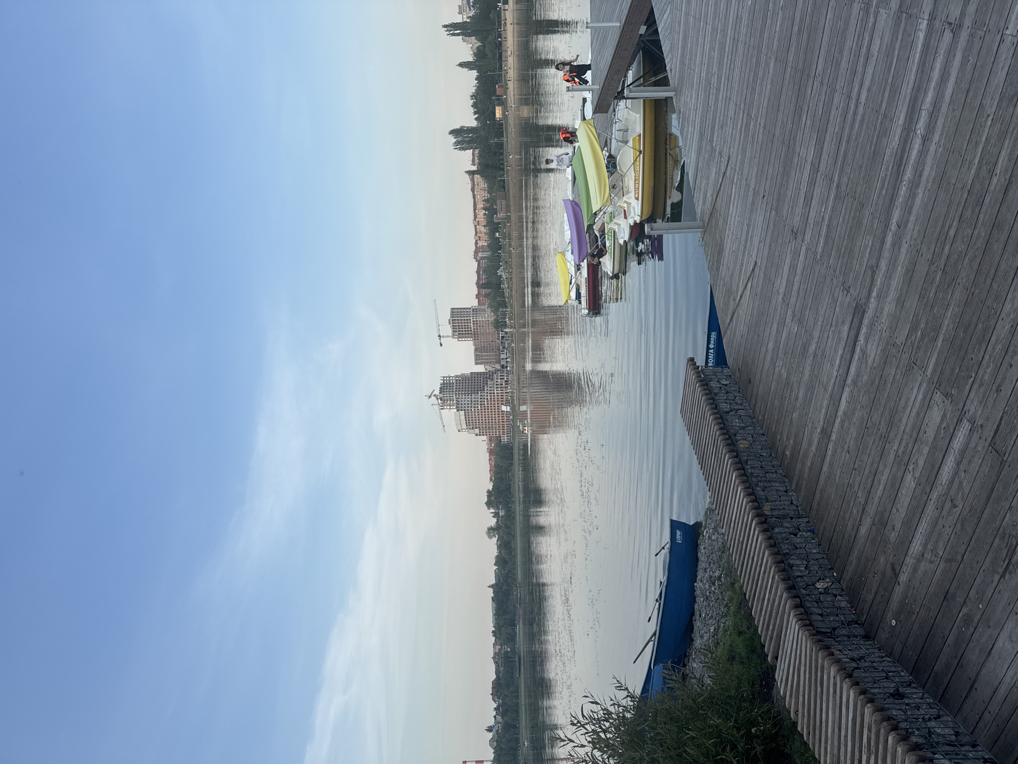 The image shows a waterfront scene in Ufa, Russia, likely during the late afternoon or early evening, given the soft light. In the foreground, a weathered wooden boardwalk leads to a dock where several colorful pedal boats are moored. Some of these boats have canopies in shades of yellow, green, and purple. Two individuals, one wearing an orange life vest, are near the edge of the dock, possibly preparing to board or disembark a boat.
Further out on the calm water, a blue rowboat is pulled up to the shore, resting on pebbles and grass. Along the embankment, a curved bench constructed from gabion walls filled with stones and topped with wooden slats provides seating.
In the mid-ground, the still water of a lake or river reflects the sky and the cityscape beyond. On the opposite bank, a dense line of trees separates the water from a mix of residential buildings and more modern structures. Dominating the skyline is a large, unfinished building with visible construction cranes, suggesting ongoing development. The sky is a pale blue with wispy clouds. The overall impression is one of a recreational area within an urban setting.