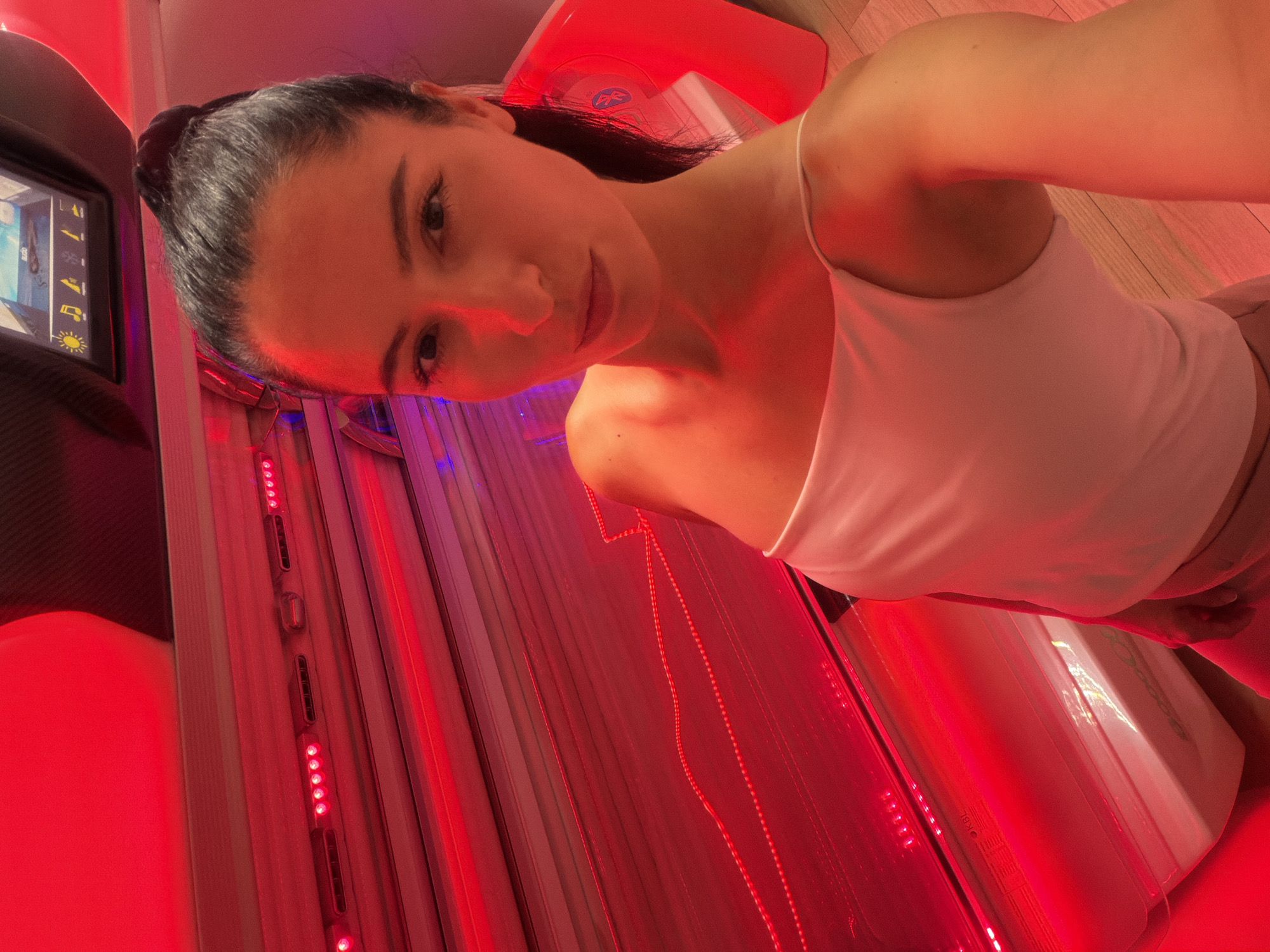 A woman with dark hair pulled into a ponytail, wearing a white tank top, is taking a selfie inside a tanning bed. The tanning bed is illuminated by red lights, and the woman's skin is reflecting the red glow. The top of the tanning bed has a control panel with a screen displaying various icons. The flooring visible in the image appears to be wooden planks. The scene appears to be an indoor commercial establishment, possibly a tanning salon.