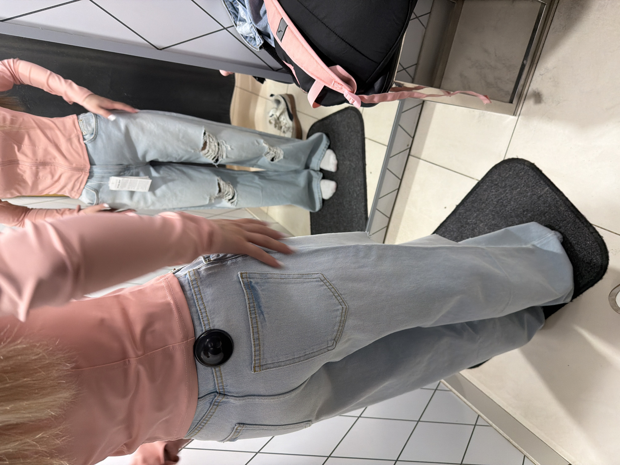 The image shows a person in a fitting room, wearing light blue jeans and a pink long-sleeved top. The person is standing on a small, dark gray mat. A mirror reflects the person and part of the fitting room. In the reflection, the person appears to be wearing ripped jeans. A black backpack with pink straps is visible in the upper right corner of the reflection. The location is most likely a clothing store, but there is no information indicating it is in Berezniki, Russia.