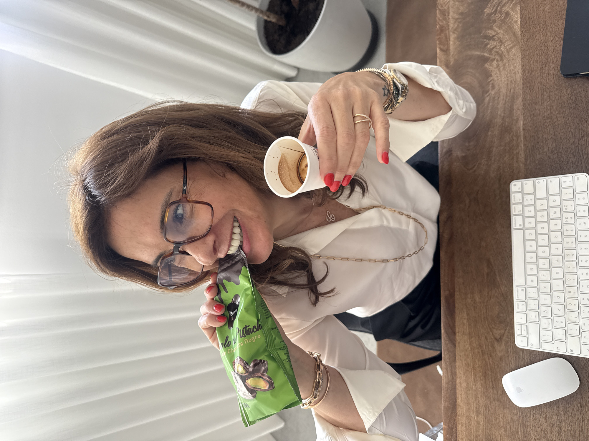 A woman with brown hair and glasses is seated at a wooden desk, facing the camera. She wears a white blouse and several bracelets on her left wrist. Her nails are painted red. She is holding a nearly empty paper coffee cup in her right hand. In her left hand, she holds a green package labeled "Pistachio Chocolate Negro" and is putting it in her mouth. A silver necklace is around her neck. On the desk to her right are a white wireless keyboard and mouse, and a dark rectangular object. In the background, there is a white plant pot with a dark plant inside. A white curtain hangs behind her. The location is not evident from the image, and there is no direct indication of Torres Vedras, Portugal.