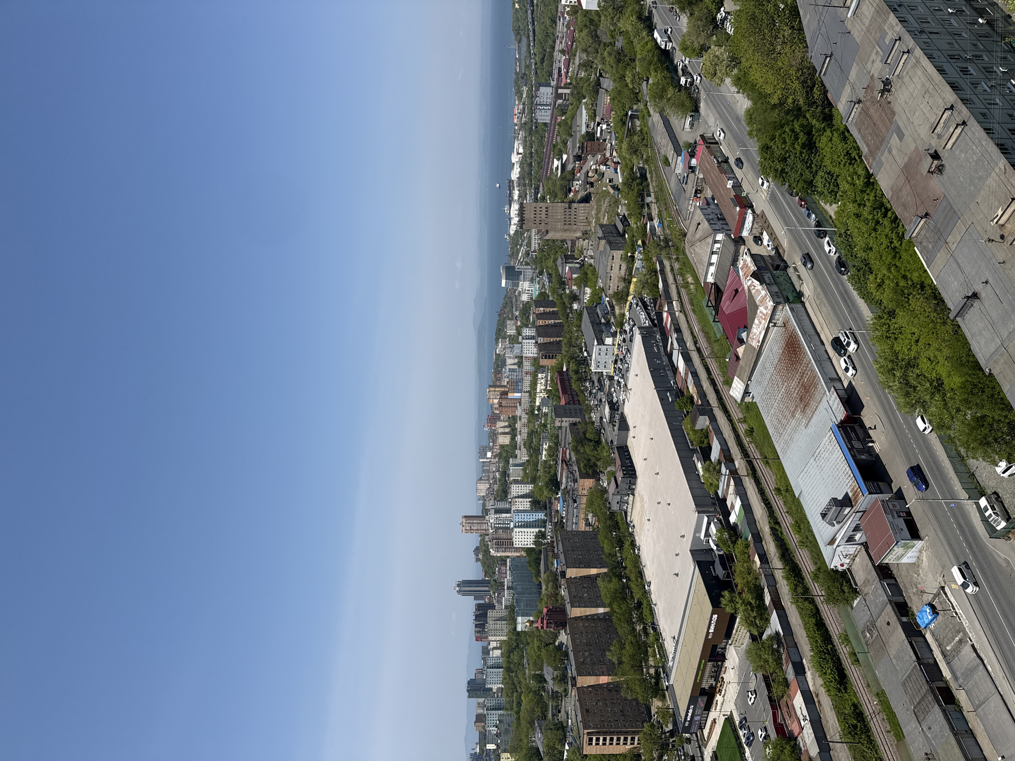 The image shows an aerial view of Vladivostok, Russia. The skyline includes buildings of varying heights, ranging from low-rise residential structures to taller commercial buildings. Vegetation is visible, including trees lining streets and scattered in urban areas. A body of water, likely the sea, forms the horizon. The perspective suggests the photo was taken from an elevated viewpoint, possibly a high-rise building or a drone. The image reveals a blend of urban infrastructure and natural features in the city. A railway line is also visible. Vehicles, particularly cars, are seen on roads. There are no people in the image.