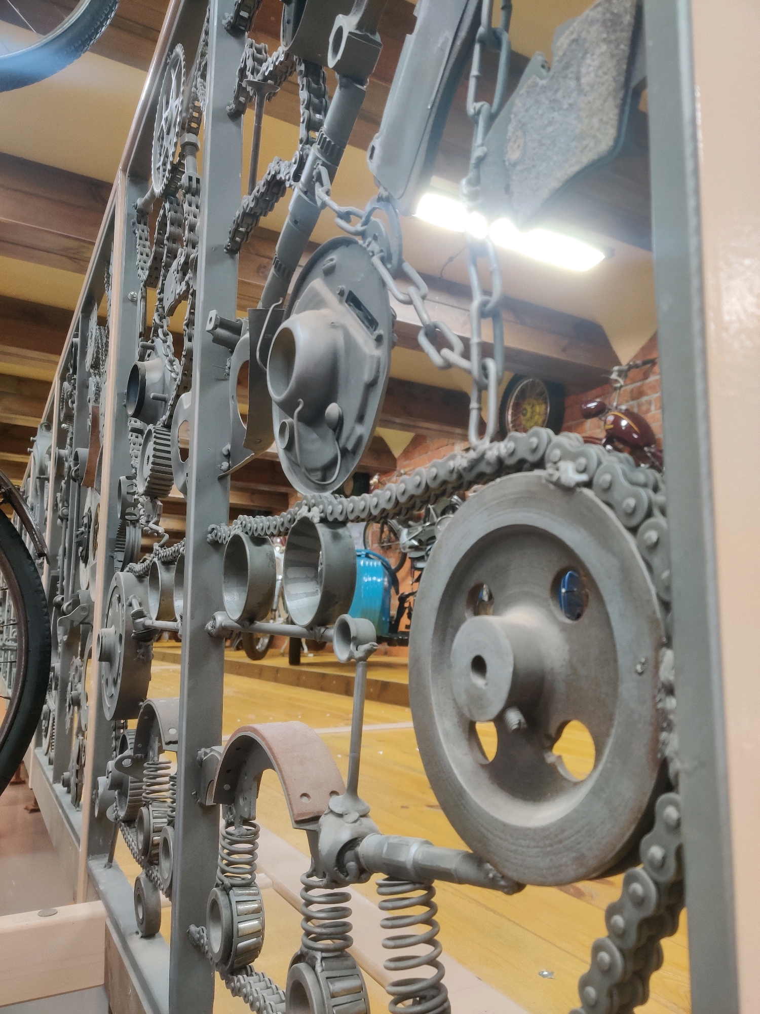 This image depicts a decorative metal fence or railing made from repurposed mechanical parts, possibly from Yekaterinburg, Russia. The structure is predominantly gray and features various components like gears, chains, springs, and cylindrical metal pieces. Some of these parts appear to be industrial or automotive in origin. The fence is viewed from a medium distance, and the background suggests an indoor setting with wooden beams on the ceiling and a yellow wooden floor. In the background, parts of motorcycles and other machinery are visible, hinting at a themed environment, perhaps a museum or workshop. There are no people visible in the image. The lighting in the scene appears to be artificial, with some bright lights causing reflections and glare. The overall impression is of a creative and industrial-style decorative element.