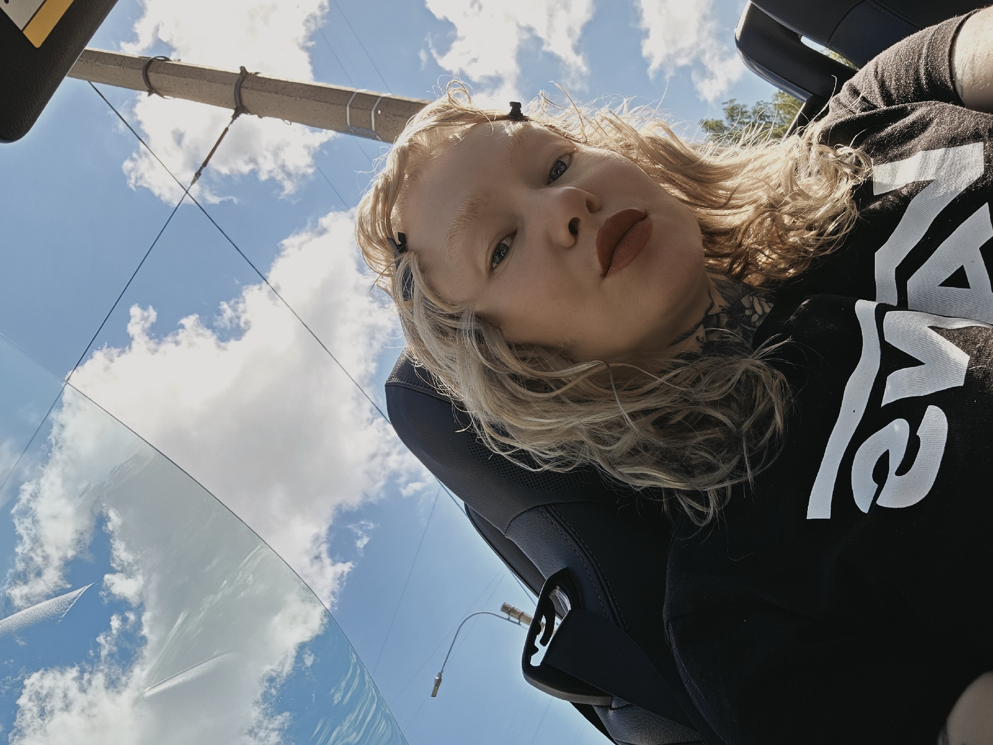 A medium shot captures a young woman with blonde, curly hair and light skin, looking directly at the camera. Her eyes are open, and she is wearing dark lipstick. She has a tattoo on her neck and upper chest. She is wearing a black t-shirt with a white graphic logo. The woman is seated in a car, indicated by the black seat and seatbelt visible in the foreground. The background shows a bright blue sky with scattered white clouds, partially reflected in the car's windshield or roof. A utility pole and a streetlight are visible in the upper right portion of the image. The image appears to be taken outdoors on a sunny day, possibly while the car is in motion given the perspective and reflections.