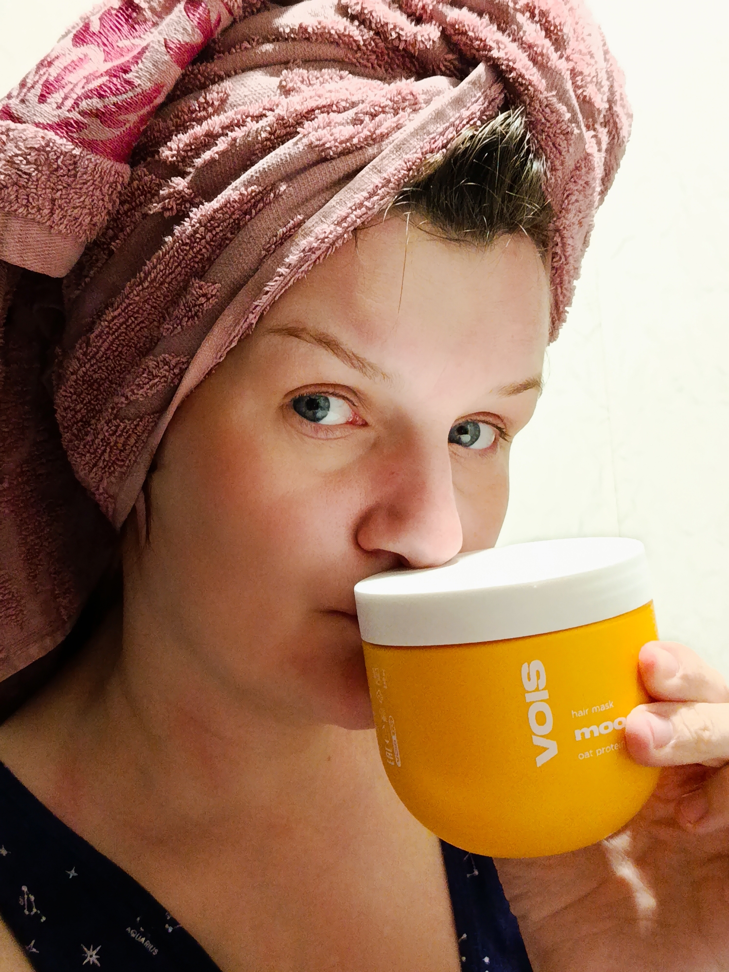 A person with a pink towel wrapped around their head is holding a yellow tub of "VOIS hair mask moo oat protein" up to their face. The person has blue eyes and is wearing a dark blue top with a constellation pattern. The background appears to be a white or light-colored wall. The location Фоки, Russia is not discernible from the image.