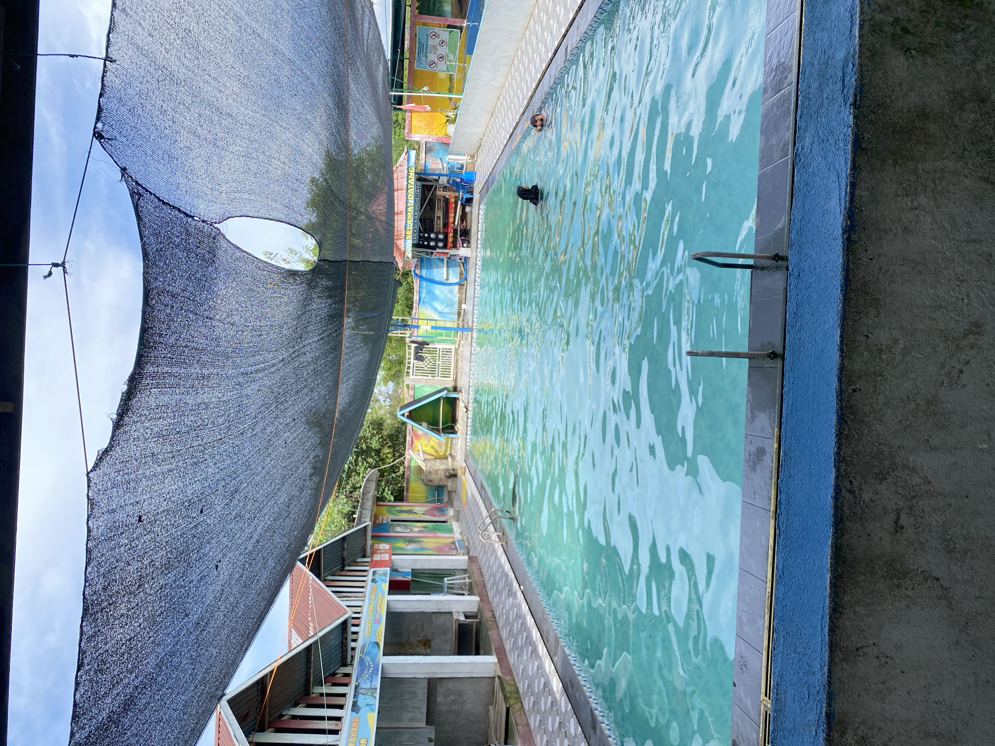 The image shows an outdoor swimming pool area. A large, dark mesh shade covers a significant portion of the scene, with a hole in the center. The pool itself is filled with clear, turquoise water and has a tiled edge. Two individuals are visible in the water, one wearing a black headscarf and the other partially submerged. To the left of the pool are several small buildings with colorful murals painted on their walls and orange-tiled roofs. A fence is visible behind the buildings. To the right of the pool, there is a small stand with a sign that reads "SELAMAT DATANG" and "KREASI KAB. PIDI JAYA", suggesting it is an entrance or information booth. Further to the right, there are more colorful walls and a white fence. The overall environment appears to be a recreational area, possibly a water park or a public swimming facility, under a partly cloudy sky.