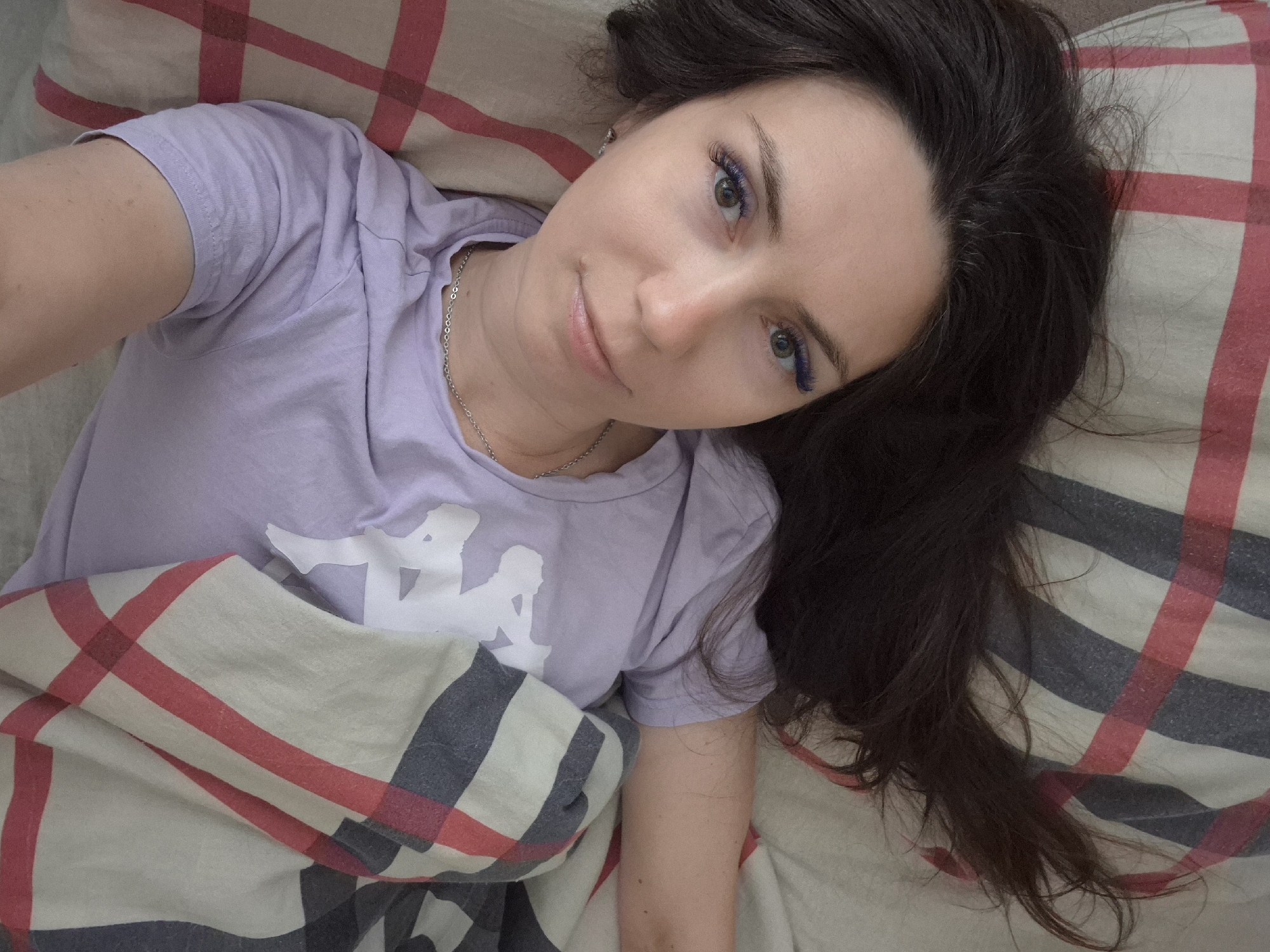 The image shows a young woman lying in bed, looking up at the camera. She has long, dark brown hair and light green eyes. Her eyelashes appear to be dyed a vibrant blue. She is wearing a light purple t-shirt with a white logo. She is partially covered by a blanket with a plaid pattern of red, beige, and dark blue stripes. The background consists of a checkered pillow and bedding. The scene is indoors, likely in a bedroom. There is no visible indication of the city of Chaikovsky, Russia, or any other specific location.