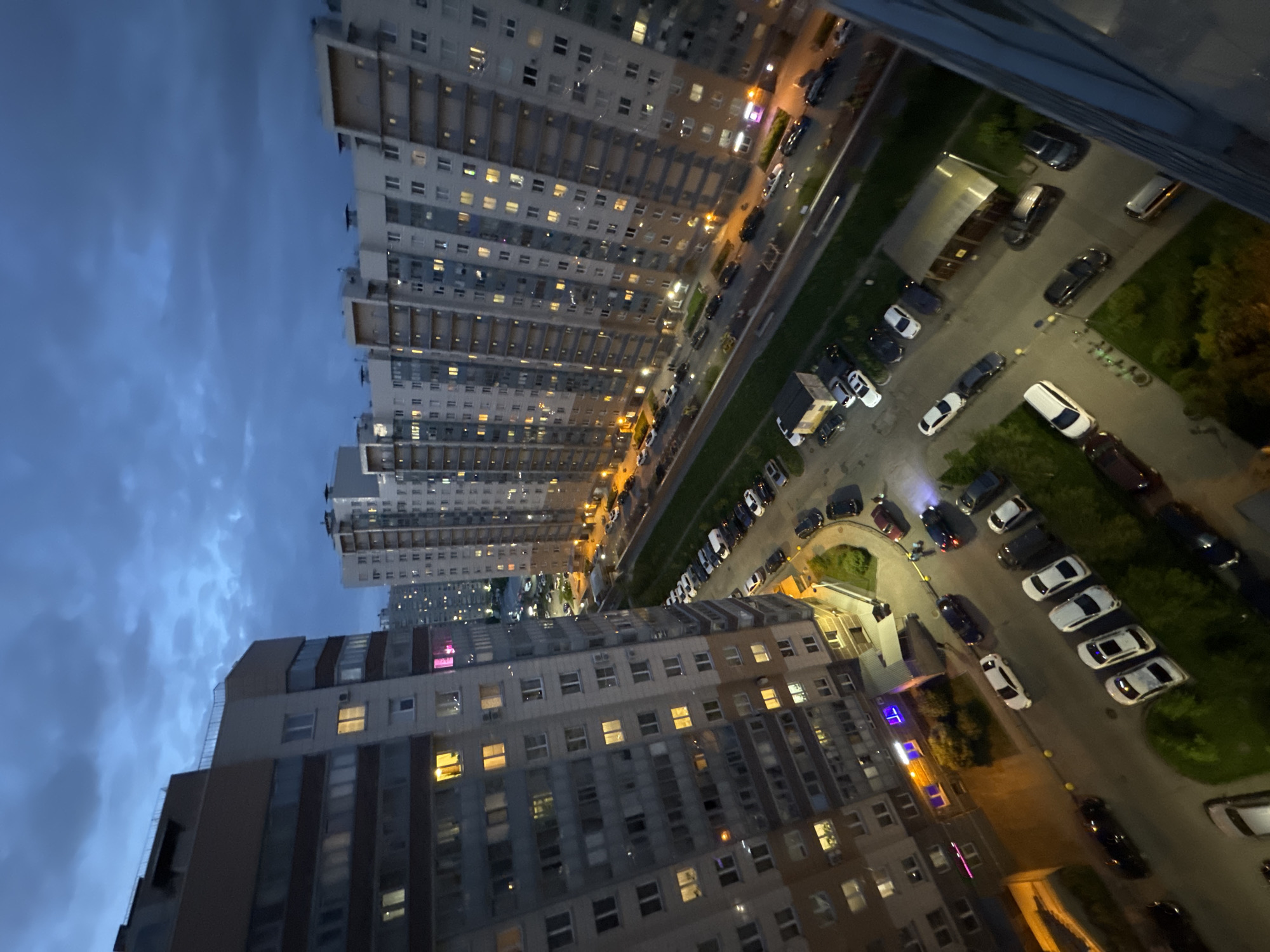 The image shows an urban scene at twilight. Several multi-story apartment buildings with lit windows dominate the view. A road lined with parked cars runs between the buildings and green spaces. The sky is a dusky blue with visible cloud formations. The overall scene appears to be taken from an elevated position within the same complex, such as a high-floor apartment.