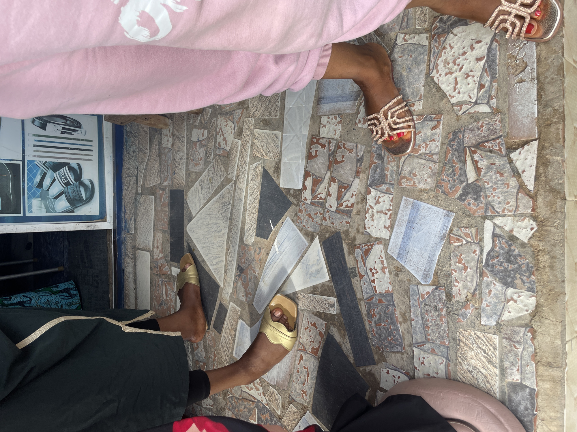 The image shows the lower bodies and feet of two people standing on a patterned tiled floor. One person is wearing a light pink garment, possibly a sweatshirt or robe, with a white graphic design on it. They are wearing rose gold sandals with an intricate, geometric strap design and red nail polish on their toes. The other person is wearing a dark green garment, possibly a dress or abaya, with gold trim. They are wearing gold slide sandals. The tiled floor is made up of irregularly shaped pieces of stone and ceramic in shades of gray, white, blue, and reddish-brown. Some of the tiles appear chipped or worn. In the background, a white cabinet or shelf unit with metal rods is visible, along with some fabric items. The lighting suggests it is daytime, and the setting appears to be outdoors or in a covered outdoor area. The overall impression is casual. There is text visible on some printed materials on the cabinet in the background, but it is not legible. The location context is Jimeta, Nigeria.