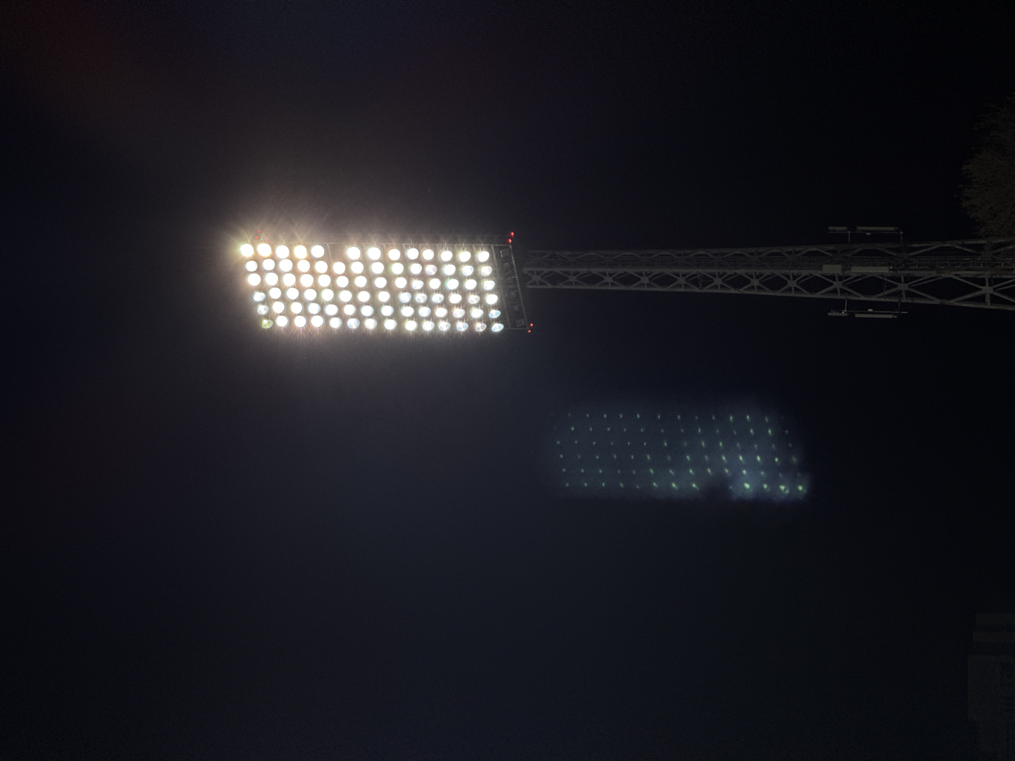 A photograph taken at night depicts a large array of lights mounted on a structure, likely stadium lights. The lights are very bright, creating a halo effect around them. Below and slightly to the right of the main light array, there's a fainter, diffuse reflection of the same light pattern. The supporting structure is visible, extending to the right of the lights. The background is a dark night sky, with some trees visible in the upper right corner. The photo was taken in Sofia, Bulgaria.
