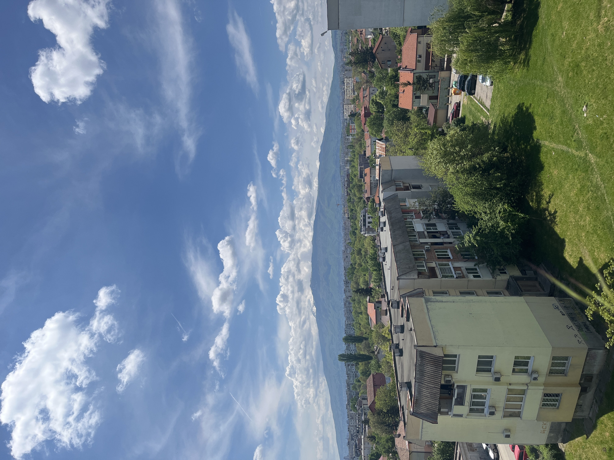 The image shows a daytime view of Sofia, Bulgaria. In the foreground, there are several apartment buildings, some with graffiti on the sides. To the right, a green lawn and trees are visible. In the middle ground, there is a cityscape with more buildings and trees. In the background, a mountain range stretches across the horizon. The sky is bright blue with scattered white clouds.