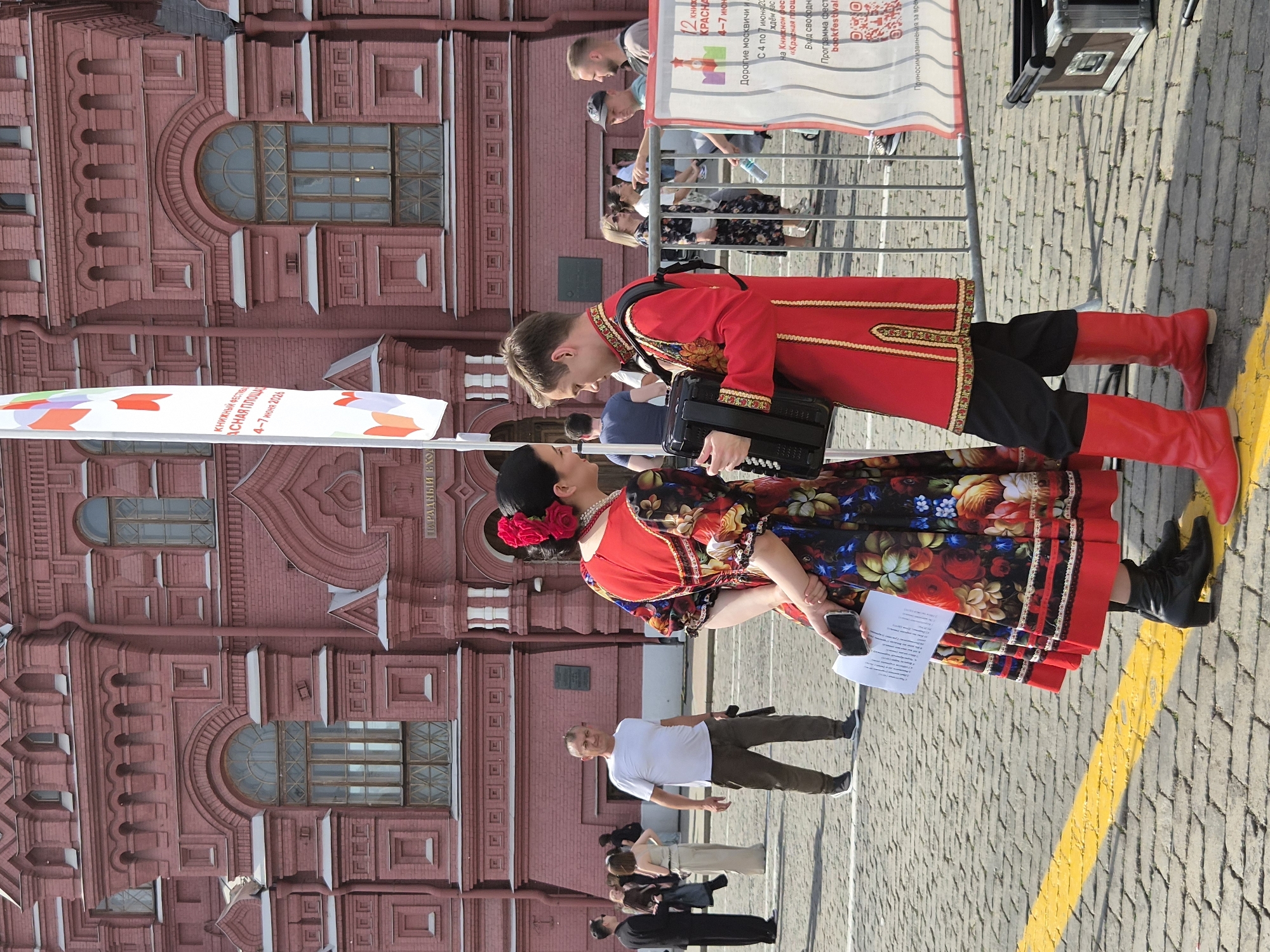 The image captures a public scene in Red Square, Moscow, Russia, against the backdrop of the State Historical Museum's red brick facade. The setting appears to be an event, identified by a partially visible white banner as the "Book Festival Red Square 4-7 June 2026".
In the foreground, two individuals in traditional Russian attire are central. On the left, a woman wears a long, dark-colored dress with vibrant floral patterns in red, yellow, blue, and green, accented with red trim at the hem and sleeves. She has dark hair styled with a prominent red flower arrangement, a pearl necklace, and wears black mid-heel boots. Her left hand holds a black smartphone and a white piece of paper. She is positioned slightly turned from the camera, facing the man.
On the right, a man wears a red tunic adorned with intricate gold embroidery on the collar, cuffs, and front opening, paired with black trousers and bright red knee-high boots. He holds a black accordion with both hands, looking towards the woman.
The ground is paved with light-colored cobblestones, bisected by a bright yellow painted line in the lower foreground. In the mid-ground, several other people are visible: a man in a white t-shirt and dark pants stands behind the main figures, looking towards the camera. Further back on the right, a group of people stands behind silver metal crowd control barriers. A black equipment case rests on the cobblestones at the far right. The scene is outdoors under bright daylight.