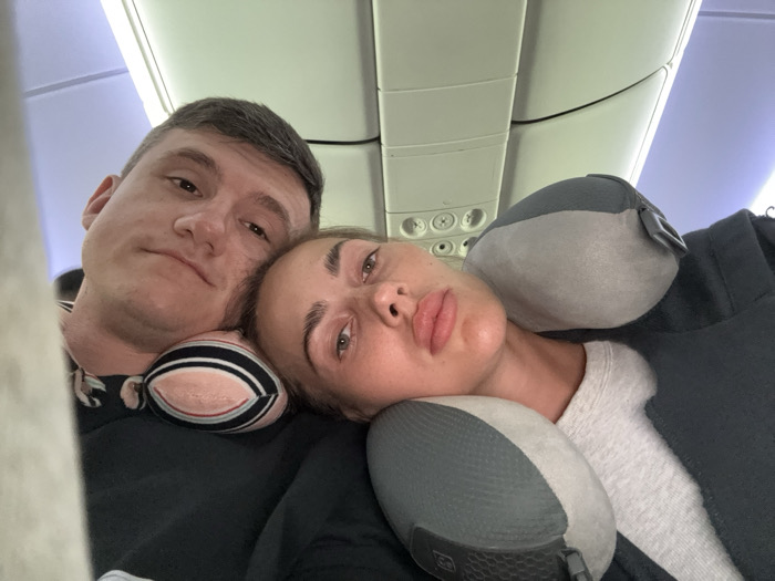 A selfie captures two individuals on an airplane. The man, positioned on the left, has short hair and wears headphones. The woman, resting her head on a grey neck pillow, appears tired or bored. They both wear casual attire. The airplane's interior, including the overhead compartment and air vents, is visible in the background. The lighting suggests daytime, potentially during a flight.