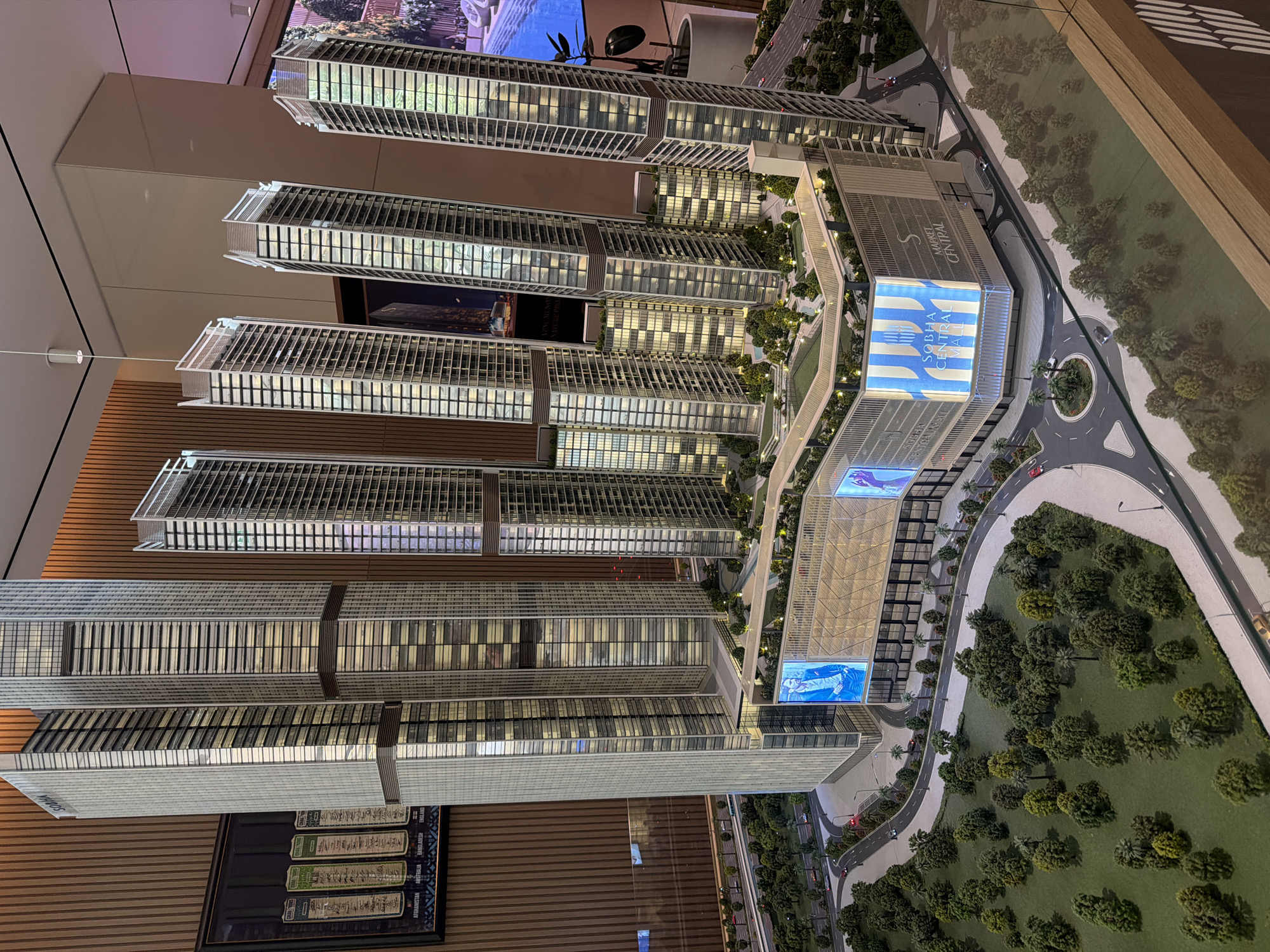 This is a photograph of a detailed architectural model of a mixed-use development. The model depicts several towering skyscrapers with a large, multi-level building at their base. The skyscrapers are designed with a modern aesthetic, featuring numerous windows and varied architectural elements. The lower building appears to be a shopping mall, indicated by signage reading "SOBHA CENTRAL MALL" and "MARKET CENTRAL." The mall has illuminated facades displaying advertisements and graphics.
The model includes a miniature landscape with trees, roads, and small vehicles, simulating a city environment. In the background, a wall with vertical wooden paneling is visible, and on the left side, a framed artwork or poster depicting various flags and country names (Afghanistan, Australia, Bangladesh, England) is displayed. The lighting in the scene suggests an indoor display. The architectural style and the presence of "SOBHA" branding suggest this model is located in Dubai, United Arab Emirates, as SOBHA is a well-known real estate developer in the region. The scale and detail of the model indicate it is likely a presentation for a property development project.