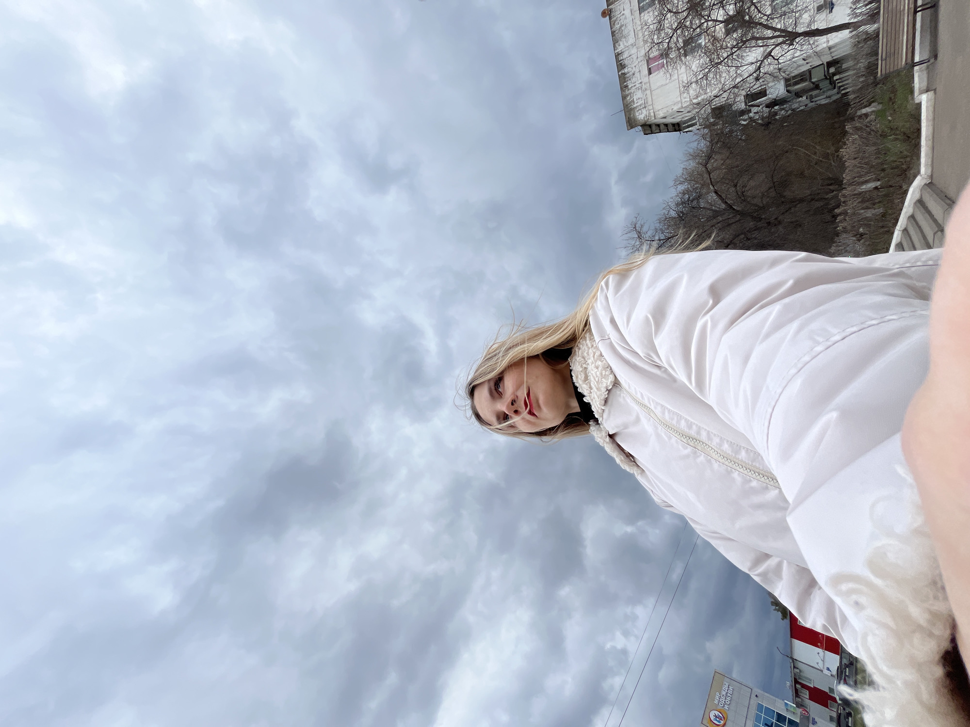 A low-angle shot shows a woman with blonde hair wearing a white jacket. The background includes a cloudy sky and some urban structures. The image appears to have been taken in Chaikovsky, Russia, as evidenced by the presence of signage in Russian. The building with the red and white stripes has signage written in Russian.