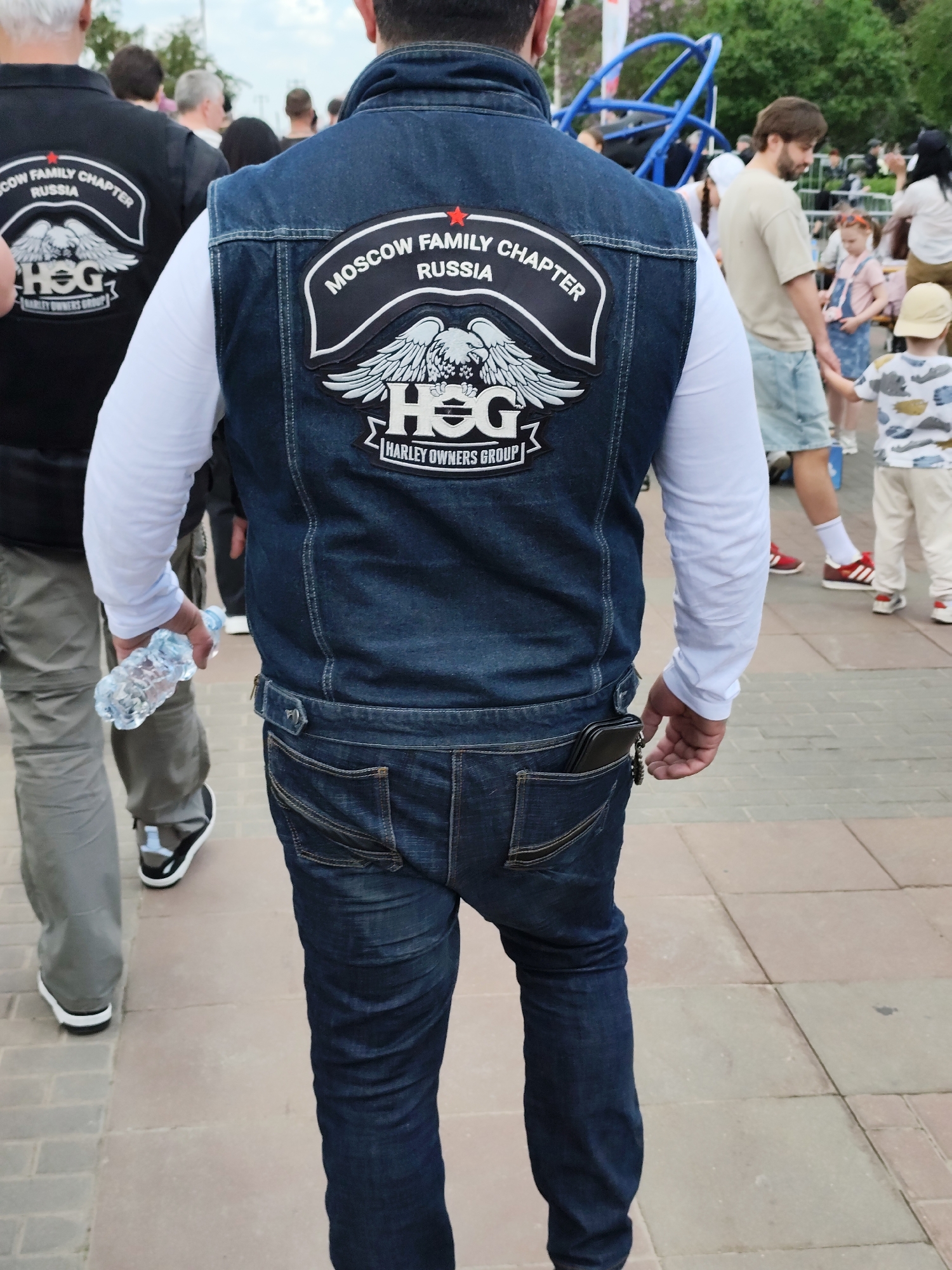 The image shows a man wearing a denim vest with a patch that reads "Moscow Family Chapter Russia Harley Owners Group." The patch features a stylized eagle design. He is wearing a white long-sleeved shirt underneath the vest and dark blue jeans. In his left hand, he holds a clear plastic water bottle. A phone or wallet is visible in his right rear pocket. Another man is standing to the left, wearing a black shirt with a similar patch. In the background, there are other people, including a child in a patterned shirt and a man in a light-colored shirt and shorts. A blue playground structure is visible further in the background. The scene appears to be in a public outdoor area. The exact location is unknown, but the presence of the Moscow Family Chapter patch suggests it may be in or associated with Russia.