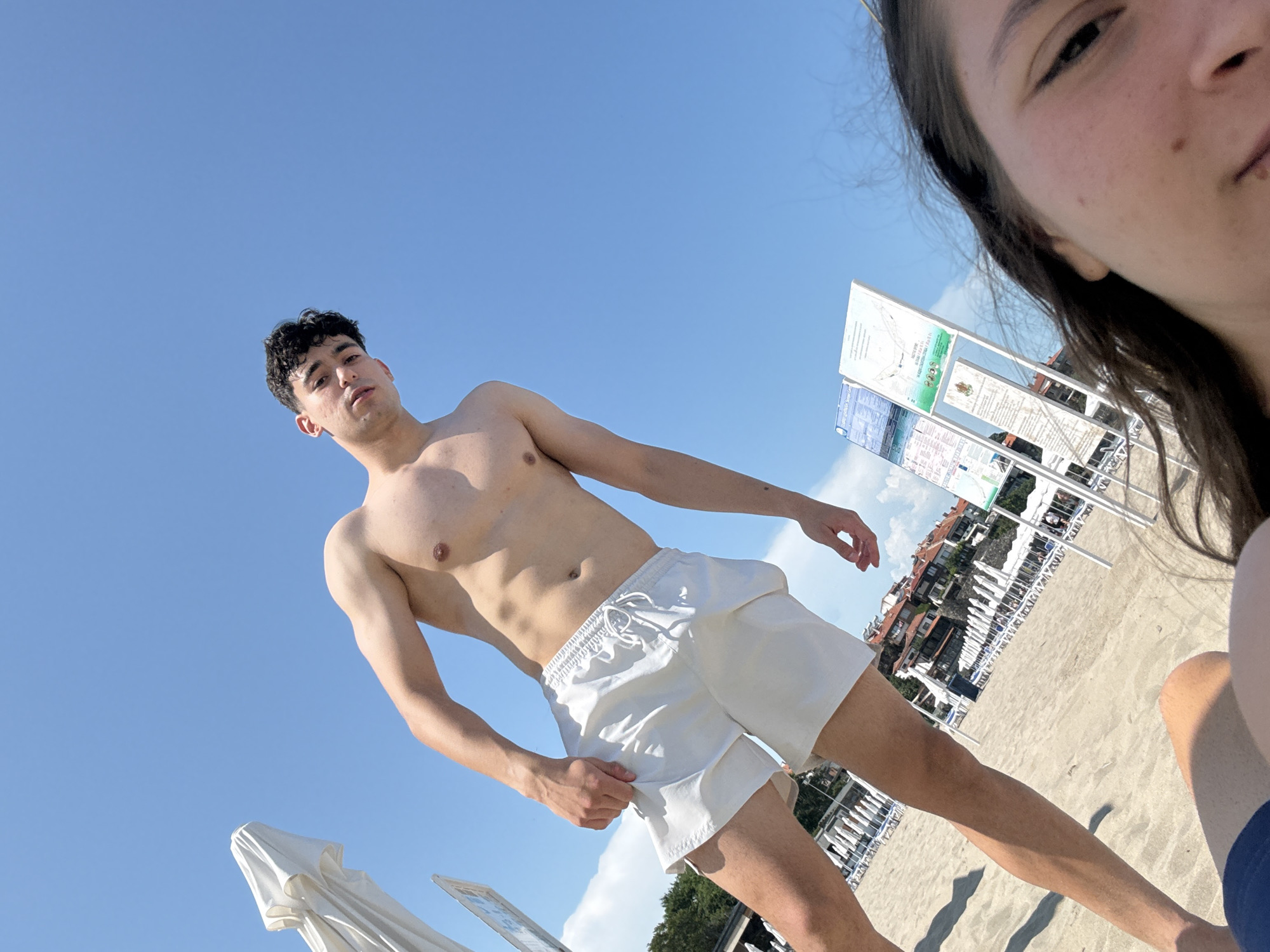 The image presents a low-angle view of a beach scene, likely in Sozopol, Bulgaria, under a clear, bright blue sky. A young man, the main subject, stands centrally, looking towards the camera. He is shirtless, displaying a toned physique, and wears white swim shorts with a visible white drawstring. His dark, curly hair frames a neutral expression.
A second person is partially visible on the right side of the frame, suggesting a selfie or a photo taken from a very low perspective, possibly while lying down. Only the right side of their face, including an eye, nose, and part of the mouth, along with some dark hair, is visible.
The setting is a sandy beach, illuminated by strong daylight, indicating it is likely midday or early afternoon. In the background, numerous white beach umbrellas and stacked beach chairs are visible, typical of a resort or public beach. Behind these amenities, a collection of buildings can be seen situated on a distant hillside.
Several information boards stand upright to the right of the man. While the detailed text on these boards is largely illegible in the main image, closer inspection from crops reveals text that appears to be in Cyrillic script, consistent with the specified location of Bulgaria. These boards likely convey information or regulations pertinent to the beach area. To the left, a folded white beach umbrella is partially visible. The overall impression is a casual, relaxed moment captured during a sunny day at the beach.