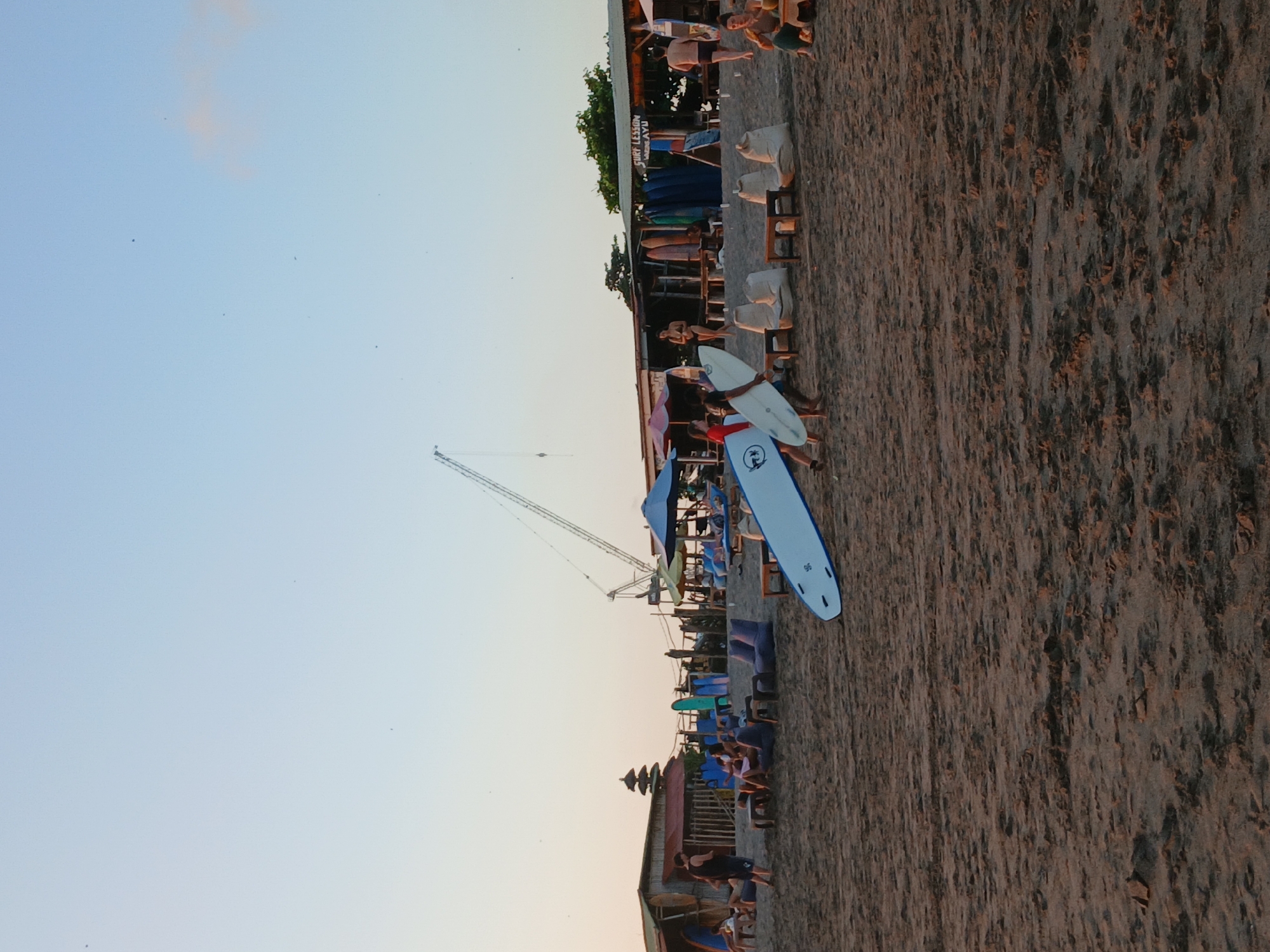 The image shows a beach scene in North Kuta, Indonesia, at dusk. The sand is dark and textured. In the foreground, a white surfboard is partially visible, lying on the sand. In the middle ground, several people are walking on the beach, some carrying surfboards. There are also beanbag chairs and low tables on the sand, as well as some surfboards leaning against a building. In the background, there are several low-rise buildings with thatched roofs, some umbrellas, and a construction crane. The sky is a pale blue with some wispy clouds. A sign on one of the buildings reads "SURF LESSON".