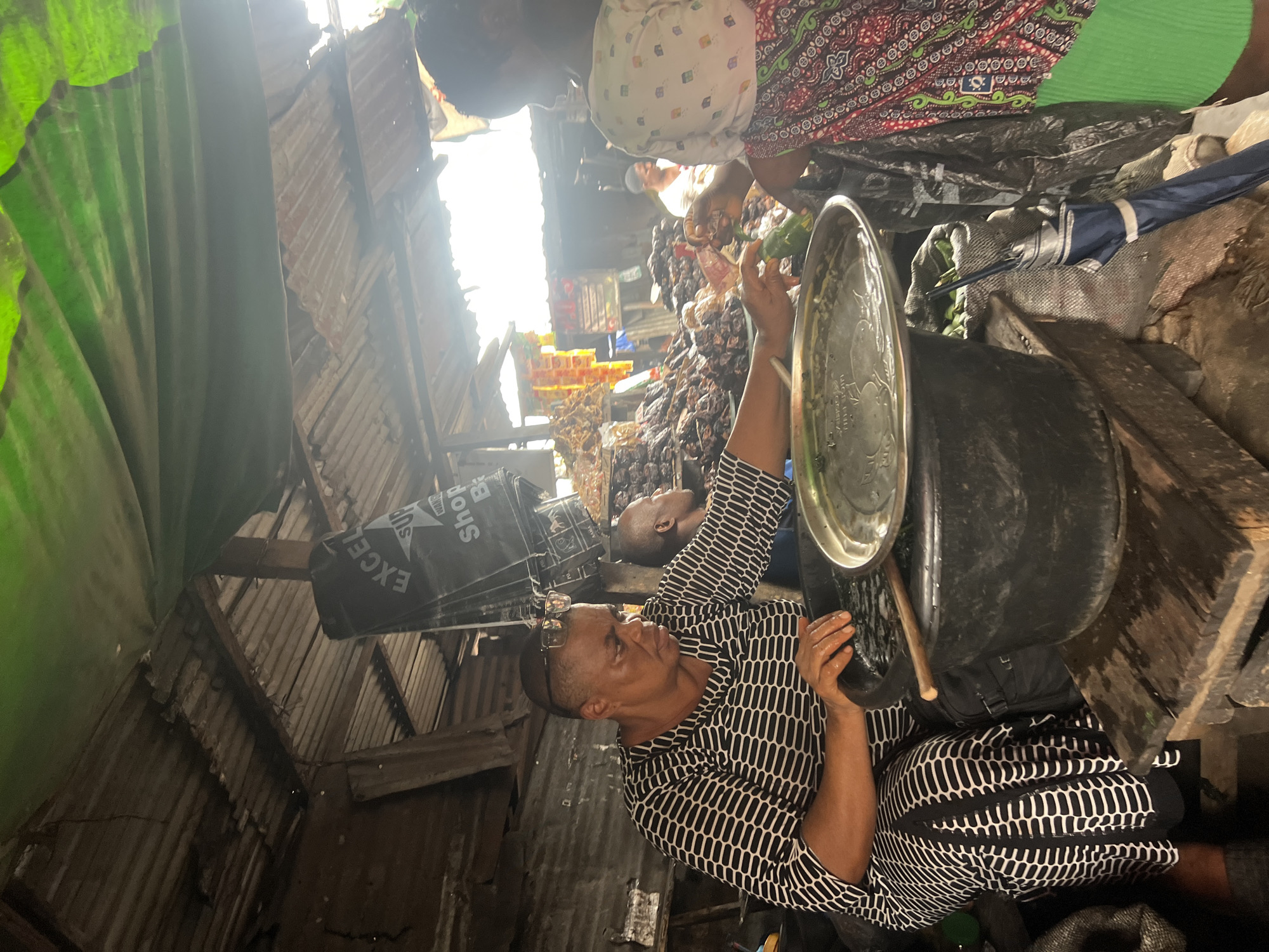 The image depicts a market scene in Nkpor, Nigeria. A woman, dressed in a black and white striped dress, is seated and appears to be preparing food. She is leaning over a large black basin filled with water, and her hands are in the basin. In front of her is a metal tray. Another woman, with her back to the camera, wearing a white top with a colorful patterned shawl and a green skirt, is interacting with the seated woman, possibly handing her something. Behind them, dried fish and other goods are piled high, indicating a marketplace. The setting appears rustic, with a corrugated metal roof and wooden structures. The overall impression is one of a bustling local market.
