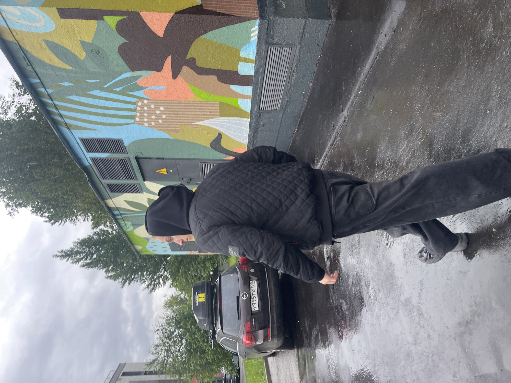 The image shows a man standing on a wet sidewalk in front of a mural and a black Opel Astra car with a roof box. The license plate on the car reads "Y 995 TX 190," indicating it is registered in the Moscow region of Russia. The man is wearing dark clothing, including a jacket with a quilted pattern and a hooded sweatshirt. The background includes trees, a building, and a cloudy sky. The mural appears to depict abstract organic shapes and patterns in various colors.