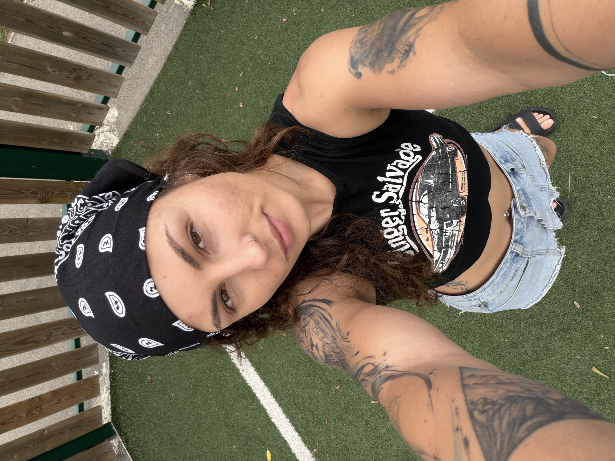 This media file is an outdoor self-portrait featuring a young adult with medium-long, curly brown hair and brown eyes, looking directly into the camera with a neutral expression. They are wearing a black bandana with white paisley patterns, a black sleeveless crop top, light blue ripped denim shorts, and black sandals.
Prominent dark, abstract tattoos are visible on both arms, with one design resembling wings or swirling patterns. A small nose piercing is also visible in the left nostril. The black crop top features white text that reads "Singer Salvage" above a graphic illustration of what appears to be a vintage vehicle or piece of industrial machinery.
The background consists of an artificial green turf surface, marked by a straight white line, suggesting a sports field or recreational area. Part of a wooden fence with green structural elements is visible on the left side of the frame. The image is brightly lit, indicating it was taken during daytime. The scene is set in an informal, outdoor environment in Kiryat Bialik, Occupied Palestine.