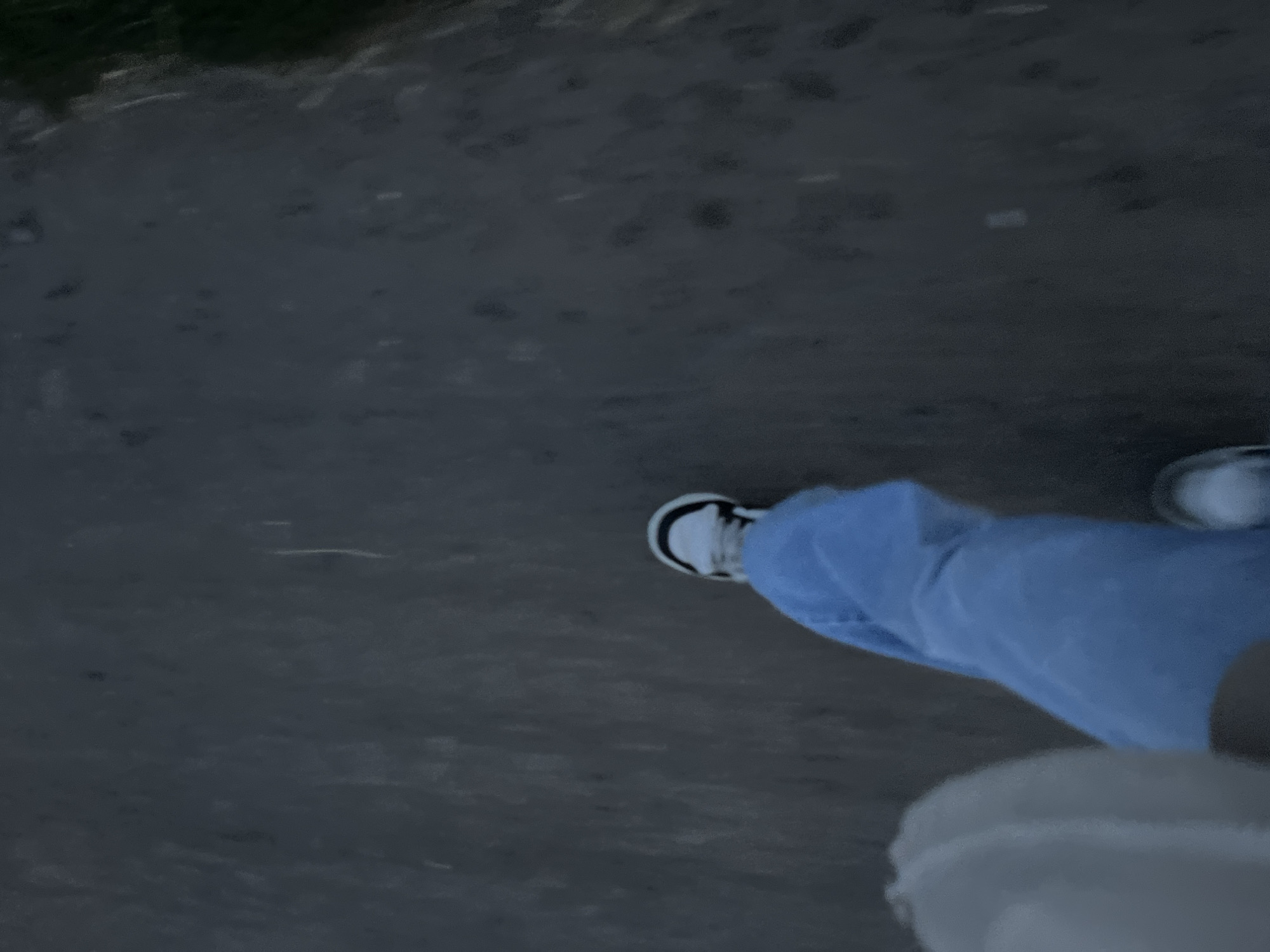 The image depicts a first-person perspective of someone walking on a dark, textured surface, possibly a dirt path or road. The lighting suggests it is either dawn, dusk, or an overcast day. The subject is wearing light-colored sneakers with black accents and loose-fitting blue pants. To the left of the frame, a portion of another person's leg, clad in light-colored clothing, is visible. The background is a blur of dark tones with some lighter streaks. There is no specific indication of the location within the city of Ustyvytsia, Ukraine.