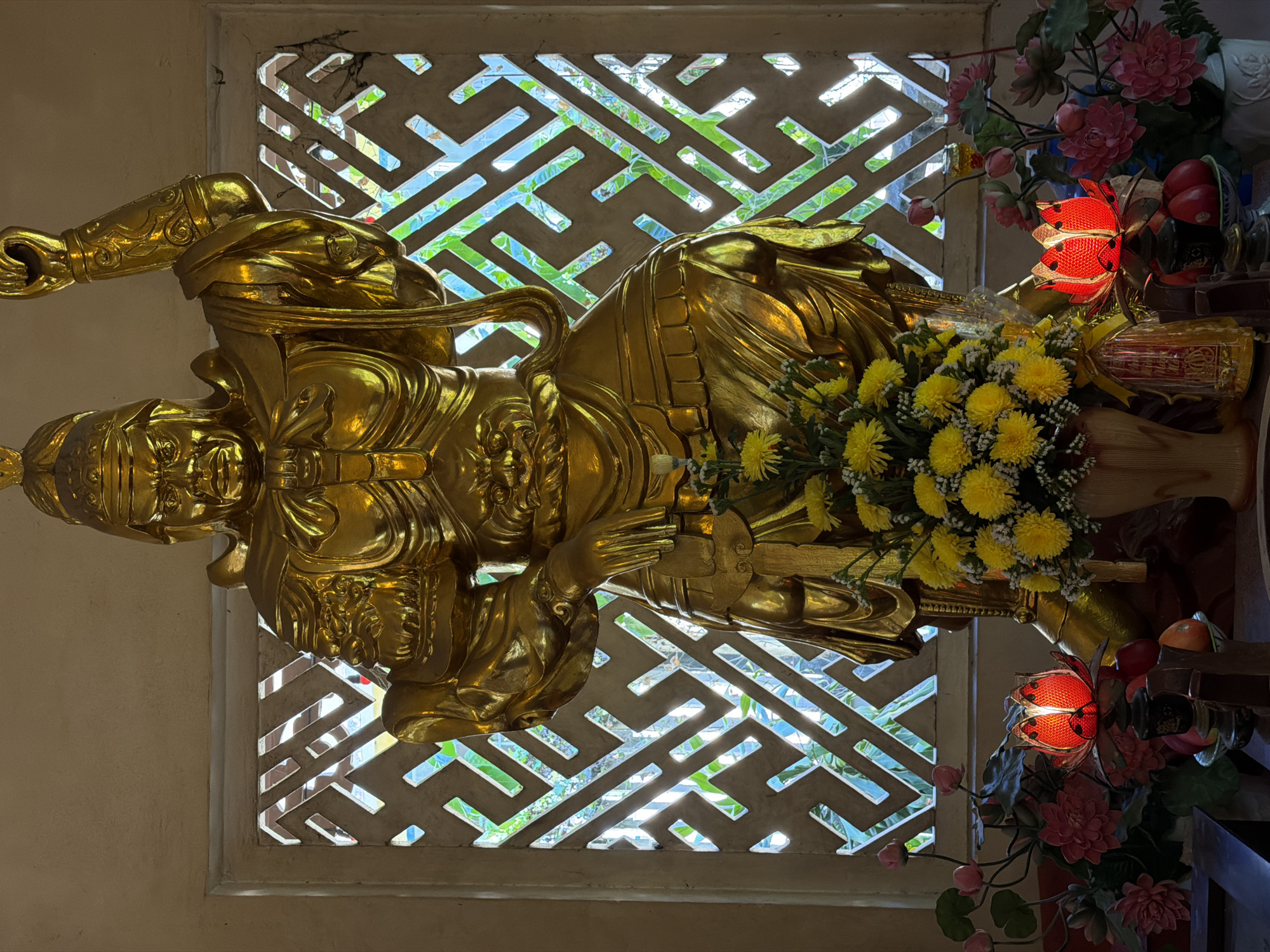 A golden, ornately detailed statue of a warrior stands in front of a geometric lattice window. The statue, positioned at an angle, is adorned with a bouquet of yellow flowers and two red lotus lanterns surrounded by pink flowers. The location is likely a temple or shrine in Nha Trang Ward, Vietnam, given the cultural and religious artifacts present.