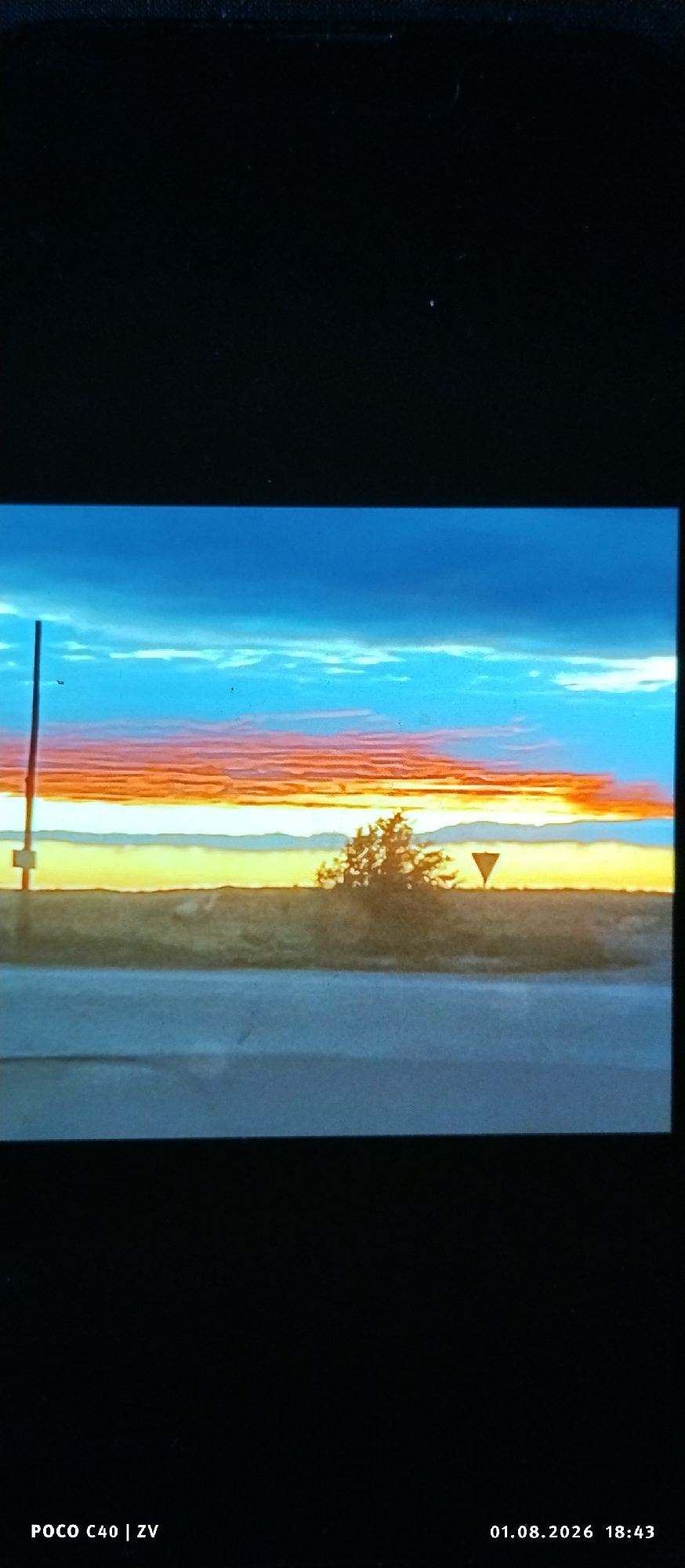 The image depicts a rural landscape at dusk or dawn, featuring a vibrant sunset. The sky displays a gradient of colors, transitioning from deep blue at the top to orange and yellow near the horizon. Wispy clouds are visible, illuminated by the setting or rising sun. A thin, vertical pole with a yellow marker is present on the left side of the frame. In the middle ground, there is a low-lying bush or small tree, and a triangular road sign is visible to its right. The foreground shows a paved surface, possibly a road, with a faint reflection or shadow. The bottom portion of the image contains text overlay indicating "POCO C40 | ZV" and a date "01.08.2026" with a time "18:43". The scene appears to be devoid of any human figures. The location is not identifiable within the city of Лубянки, Russia, based on the visual information provided.