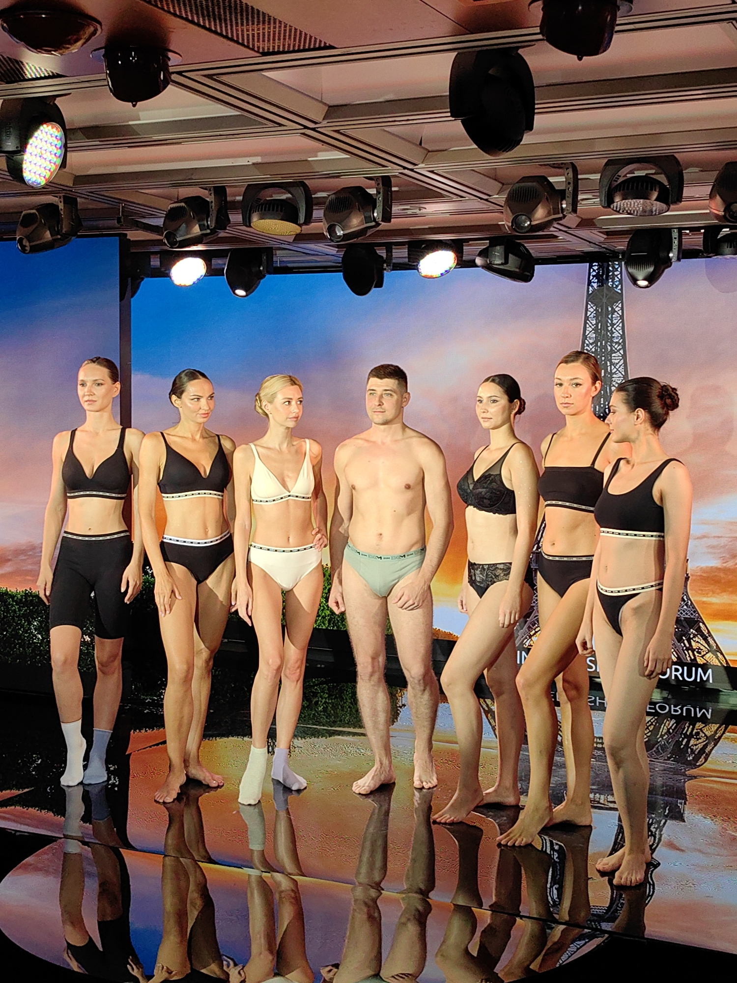 The image shows a fashion show presentation of lingerie and swimwear. Seven models, four women and three men, are standing in a line on a reflective stage. The backdrop features a sunset scene with an image of the Eiffel Tower, suggesting a Parisian theme. The models are displaying various pieces of underwear and swimwear. The lighting is provided by spotlights suspended from the ceiling. The scene appears to be an indoor event, possibly a fashion runway or product launch. There is no visual information in the image to indicate that the event is located in Moscow, Russia.