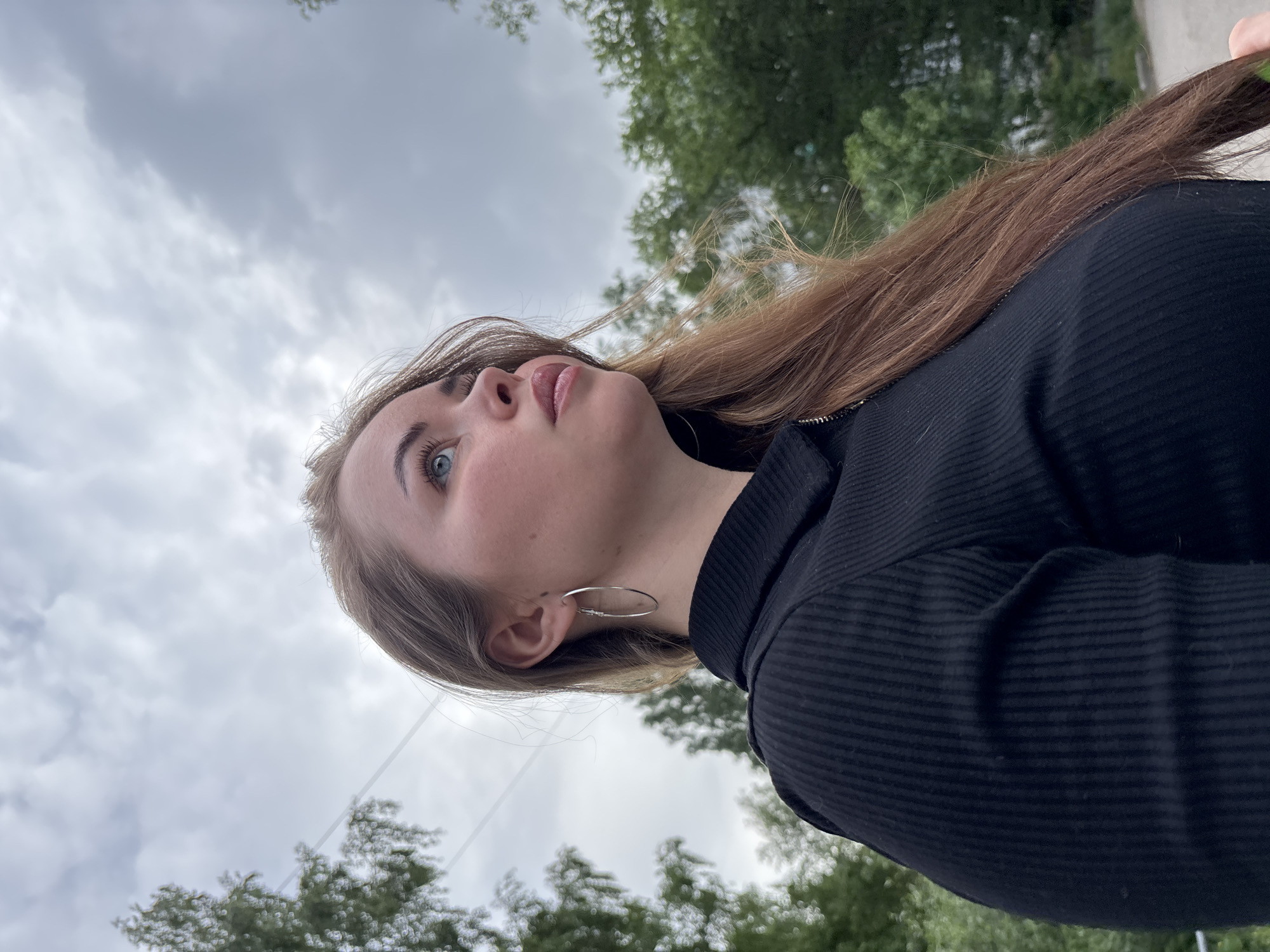 A woman with fair skin, blue eyes, and light brown hair stands outside, looking upwards. She is wearing a black ribbed top and silver hoop earrings. The background consists of a cloudy sky and green trees. The location is possibly in the city of Izhevsk, Russia.