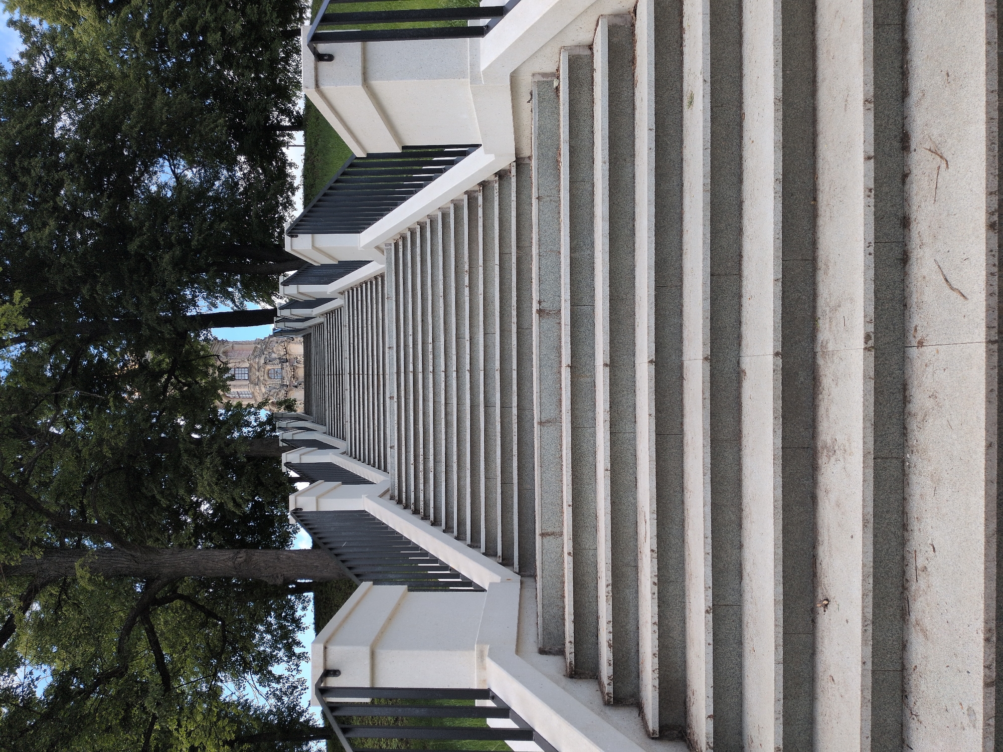 The image displays a long, wide outdoor staircase ascending upwards, flanked by railings on both sides. The steps are constructed from a light grey, speckled stone or concrete, exhibiting some natural wear and minor debris like leaves or dirt. On either side of the staircase, robust white stone or concrete balustrades with dark, possibly black, metal railings provide a symmetrical leading line.
Large, mature trees with dense green foliage frame the scene, growing on both sides of the stairs and extending into the background. Through the canopy of these trees at the top of the staircase, a multi-story building with architectural features, including windows, is partially visible in the distance. The sky, seen in glimpses through the trees, appears clear and blue.
The scene is illuminated by bright daylight, casting shadows on the steps and indicating a sunny day. No individuals are present, and no specific activity is taking place. The setting suggests a grand public space, possibly a park entrance or an approach to a significant building in Podolsk, Russia. There is no visible text in the image.