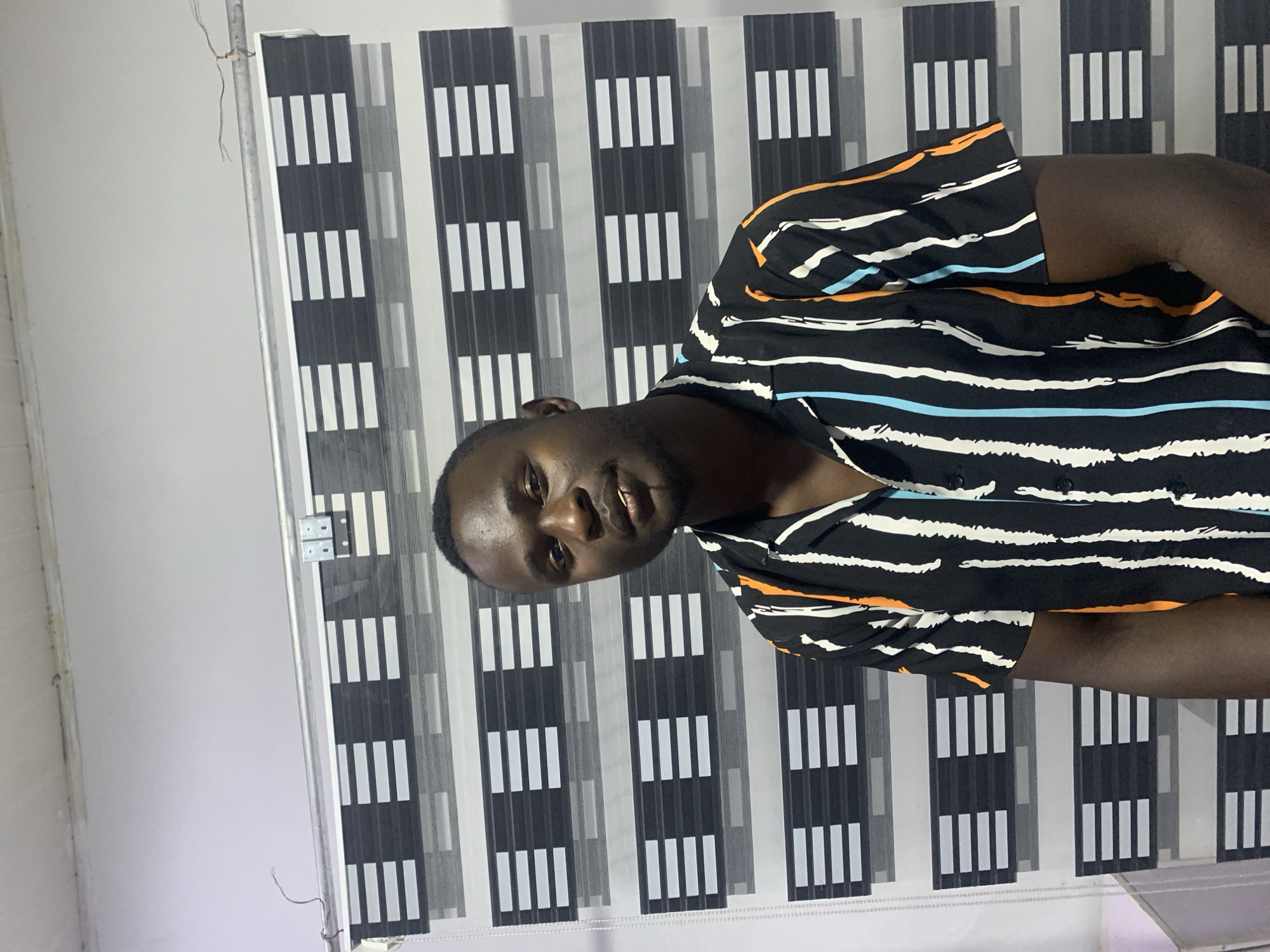 A young man with dark skin and a short beard is captured in an indoor setting, looking directly at the camera with a subtle smile. He is wearing a black short-sleeved shirt featuring a distinctive pattern of abstract, brushstroke-like designs in white, light blue, and orange.
The background behind him consists of modern window blinds, likely "zebra blinds," characterized by alternating horizontal panels. These panels are patterned with black opaque sections containing sets of four white vertical stripes, separated by translucent grey fabric, creating a repetitive and contemporary aesthetic. A metallic rod or frame, possibly part of the blind mechanism, is visible along the left side of the blinds, with a few loose wires near the top. The wall to the far left of the blinds is plain white.
The image appears to be taken indoors, suggesting an interior space in Jos, Nigeria, as per the provided context. The lighting is consistent with an indoor environment, possibly utilizing artificial light. No specific activity is taking place beyond the subject posing for the photograph. There is no visible text in the image.