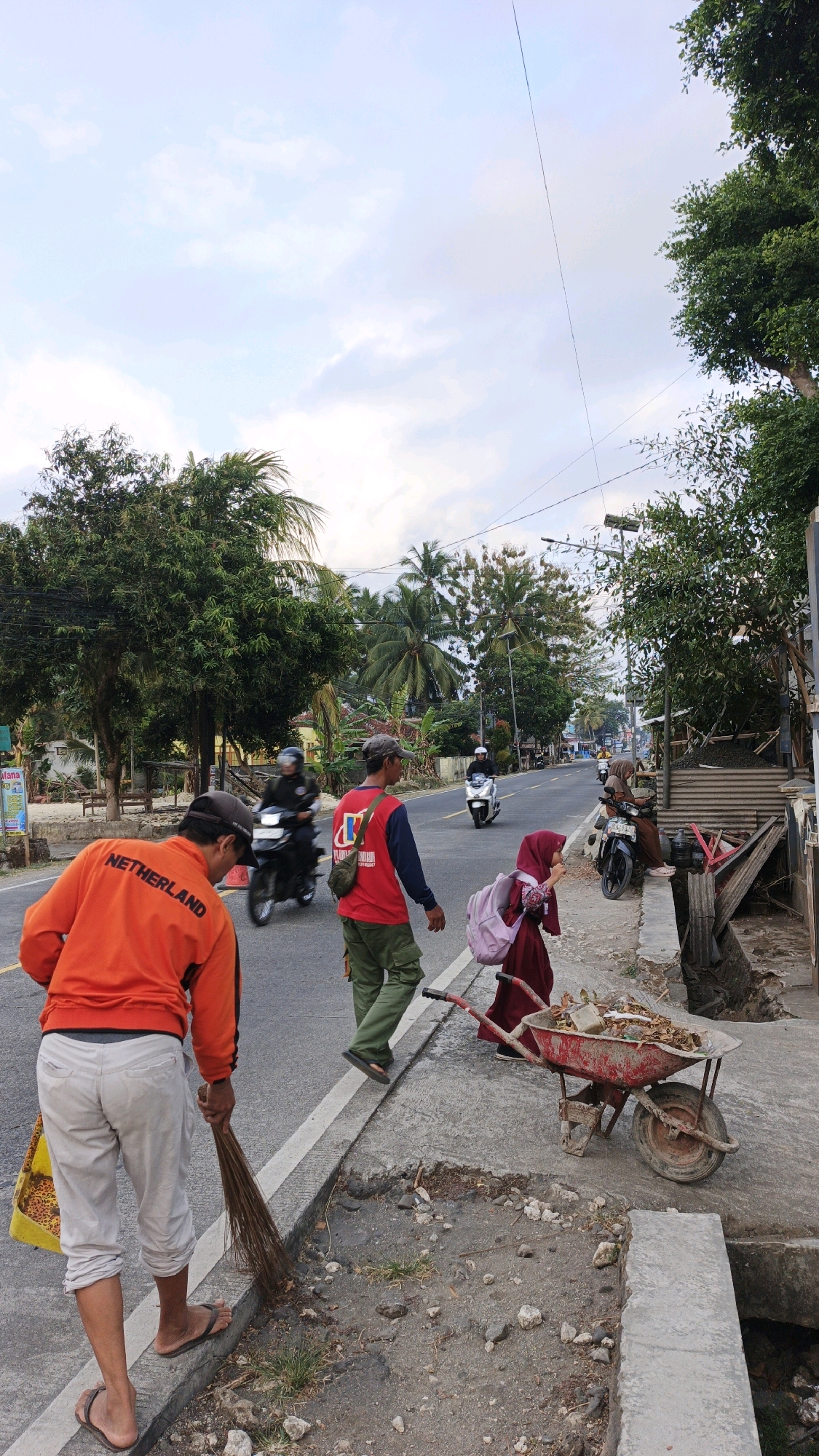 In Cibenda, Indonesia, an outdoor scene depicts a roadside with several individuals engaged in activity. The foreground shows a person wearing an orange long-sleeved shirt with "NETHERLAND" printed on the back and light-colored pants, holding a yellow bag and sweeping the road with a broom. To their right, another individual in a red shirt and green cargo pants walks along the sidewalk. Further ahead, a young person in a maroon hijab and dress carries a pink backpack, and next to them is a red wheelbarrow filled with debris. Several motorcycles are visible on the road, some with riders. The environment includes trees, a cloudy sky, and a street with a damaged sidewalk.
