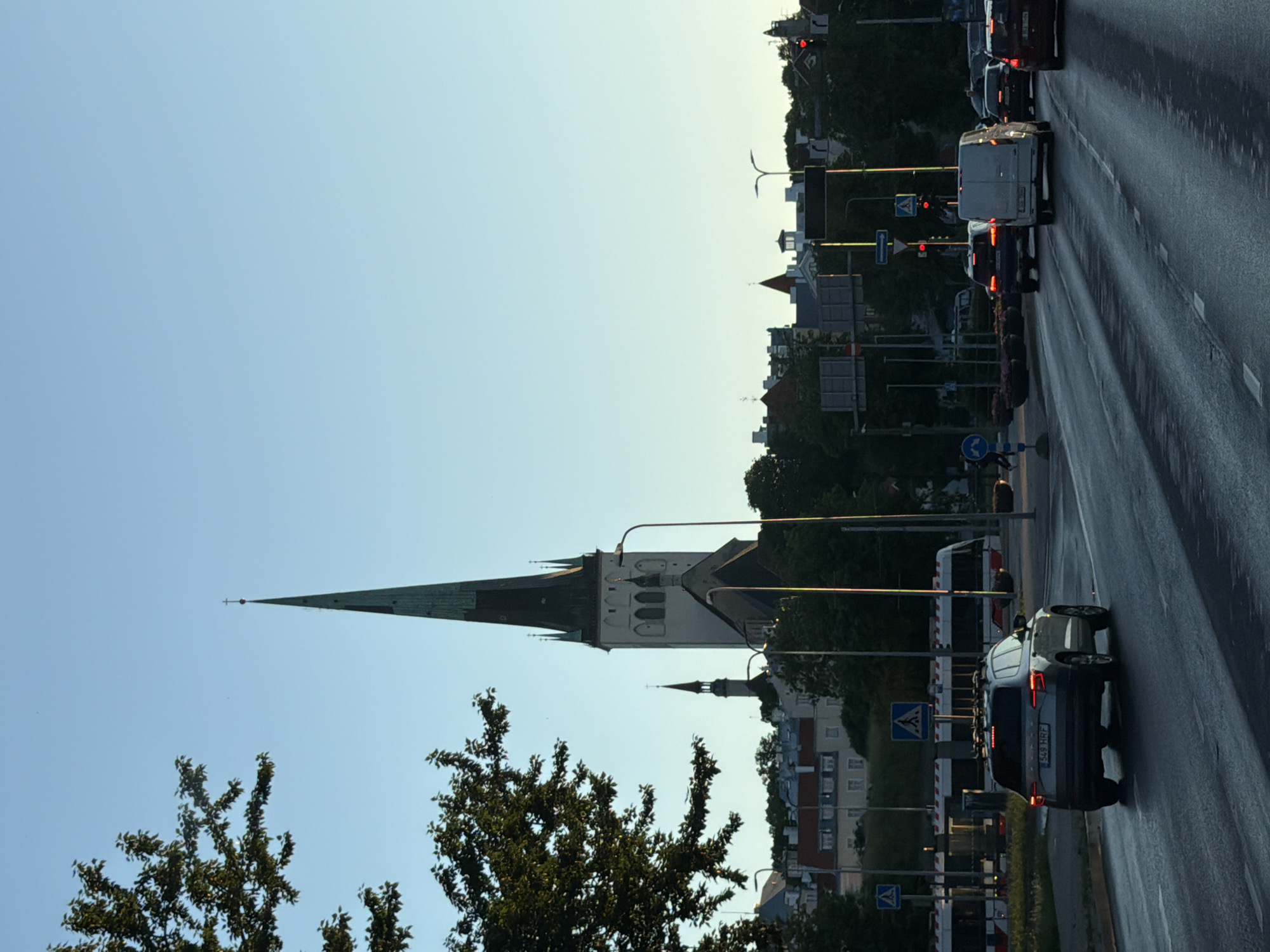 The image depicts a street scene in Tallinn, Estonia, with a prominent view of the St. Olaf's Church (Oleviste kirik) in the background. The church's tall, slender spire, topped with a cross, dominates the skyline. The sky is a clear, pale blue, suggesting it is either early morning or late afternoon. In the foreground, a wet asphalt road reflects the light, indicating recent rain or dew. Several vehicles are visible on the road, including a grey SUV with a license plate starting with "549 HRF" in the left lane, a white van, and other cars further down the road. A tram with red and white stripes is stopped or moving slowly in the background. Pedestrian crossings and traffic lights are present, suggesting an urban intersection. Trees with green foliage line the sides of the road. The architecture in the background appears to be a mix of historical buildings with red roofs and more modern structures. The overall atmosphere is that of a quiet city street during a time of day with diffused sunlight.