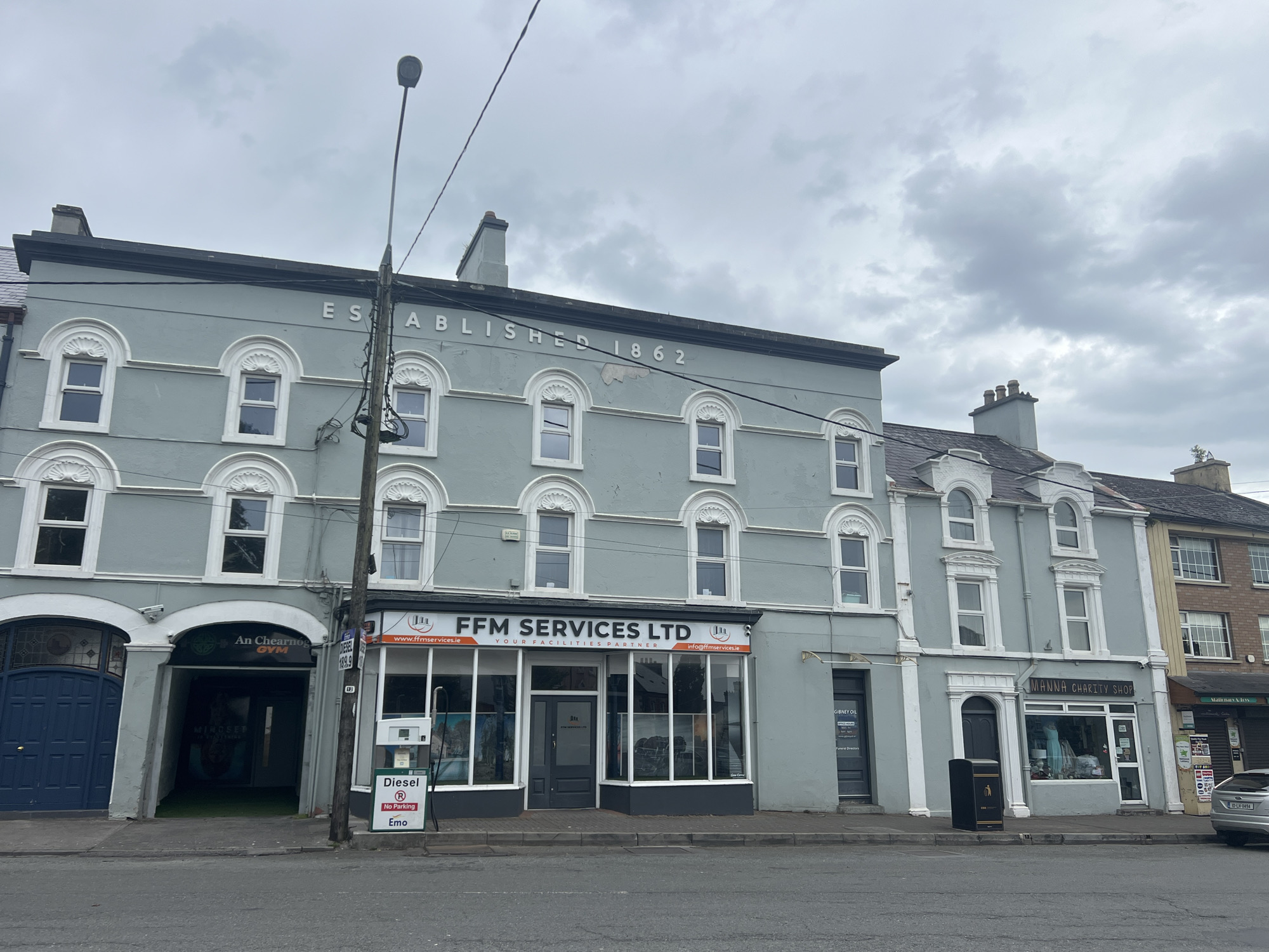 The image shows a street scene in a town with a cloudy sky. On the left is a light blue building with "ESTABLISHED 1862" written in white above the second-story windows. The building has arched windows on the second story and rectangular windows on the third story. A dark blue door is visible on the far left, leading into what appears to be a gym named "An Chearnog GYM". Next to this is a storefront with a dark gray door and large windows displaying images of machinery. Above the windows, the text "FFM SERVICES LTD YOUR FACILITIES PARTNER" is visible. A diesel pump with a "No Parking" sign is located to the right of the gym entrance. To the right of FFM Services is a building with "MANNA CHARITY SHOP" above its storefront window. A dark gray trash bin is situated in front of the charity shop. Further to the right, another building with multiple windows and an awning is visible. The street in front of the buildings is paved and appears to be a public road. A silver car is parked on the right side of the street. A lamppost with wires attached is positioned in front of the buildings.