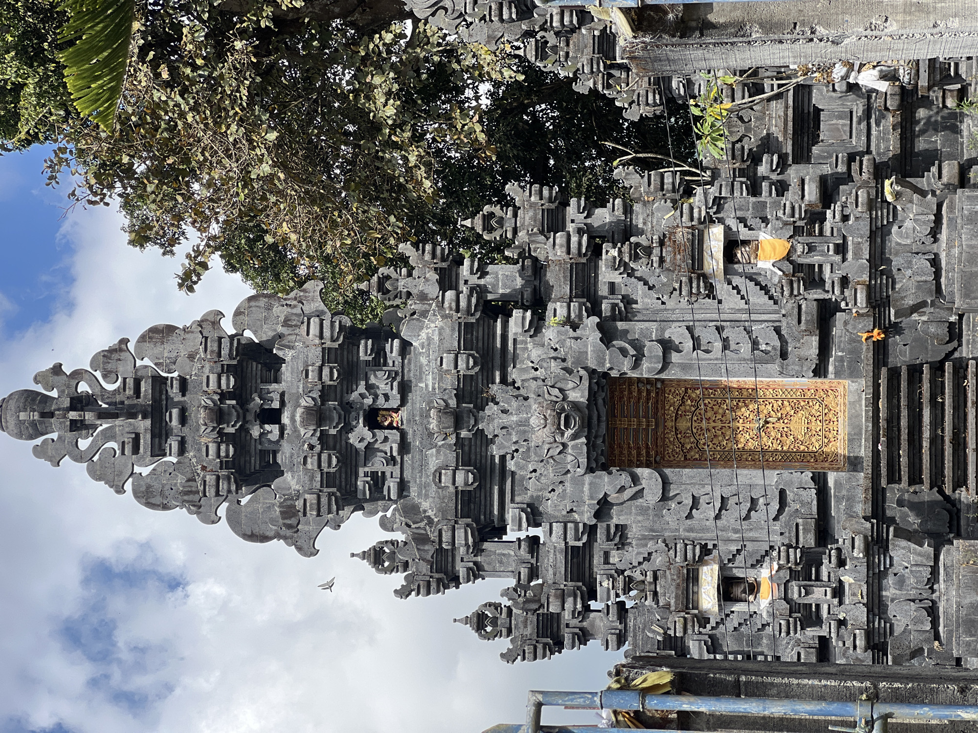 The image shows a detailed, ornate stone temple structure, possibly a Balinese Hindu temple, located in Kintamani, Indonesia. The temple is constructed from dark grey stone with intricate carvings. The central facade features a large, golden, intricately carved door. Above the door, a monstrous face is carved into the stone. The structure is tiered, with smaller decorative elements and niches on multiple levels. A small statue is visible in a niche above the door, adorned with orange and white fabric. There are steps leading up to the entrance. To the right of the temple, lush green trees with dense foliage are visible against a blue sky with scattered white clouds. The environment appears to be outdoors, under daylight.
