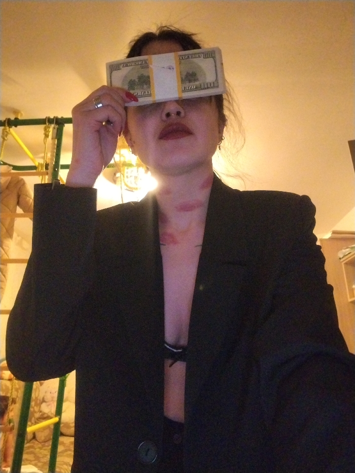 A woman in a black blazer strikes a pose, covering her eyes with a stack of hundred-dollar bills. Her lips are painted a deep red. She sports a nose ring. A few red lipstick marks are visible on her neck and upper chest. A black bra is partially visible beneath the blazer. In the background, a climbing frame and other furniture are partially visible.